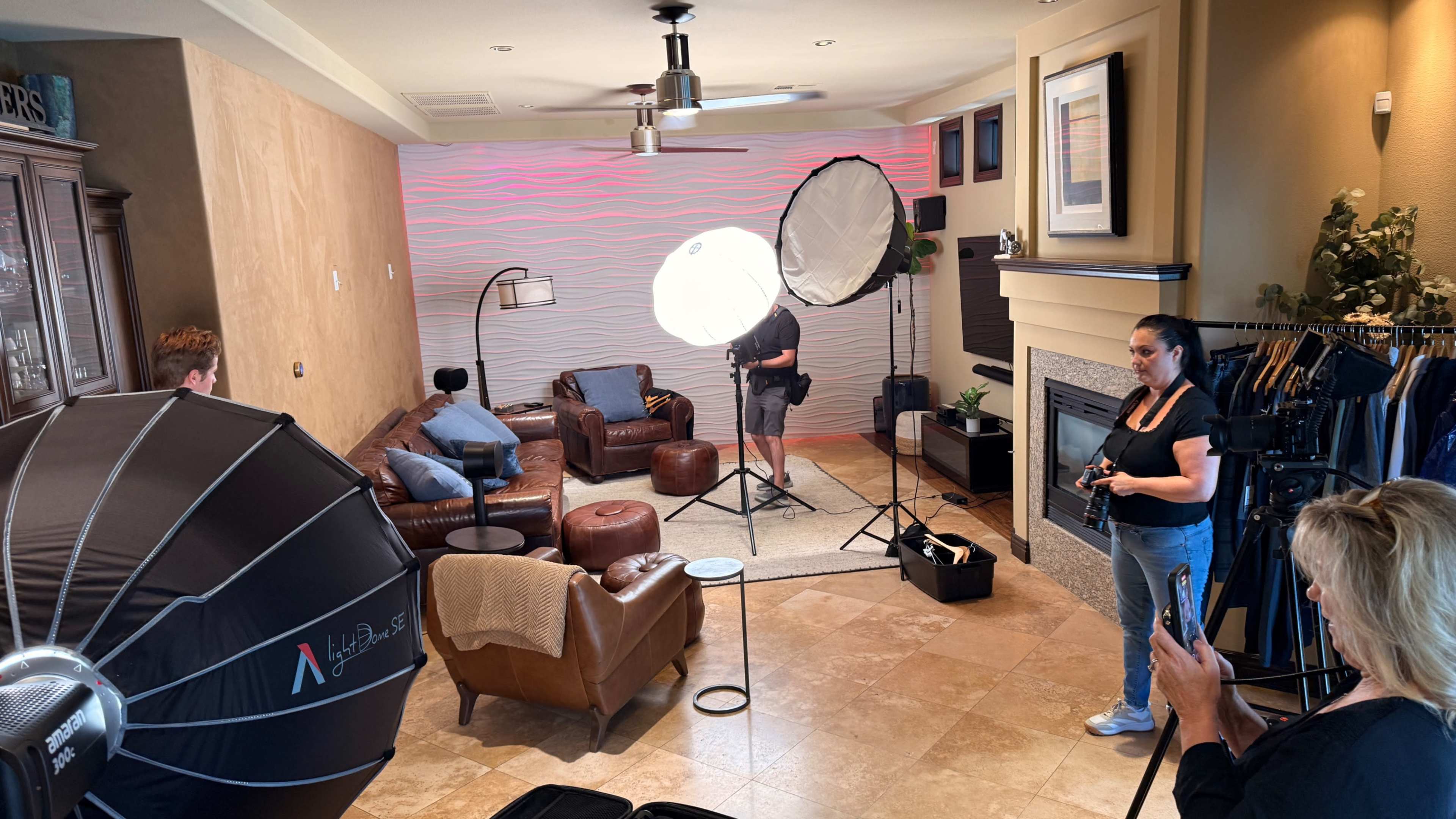 A photography session is taking place in a well-lit living room, featuring soft lighting equipment and a photographer directing a model near a couch.