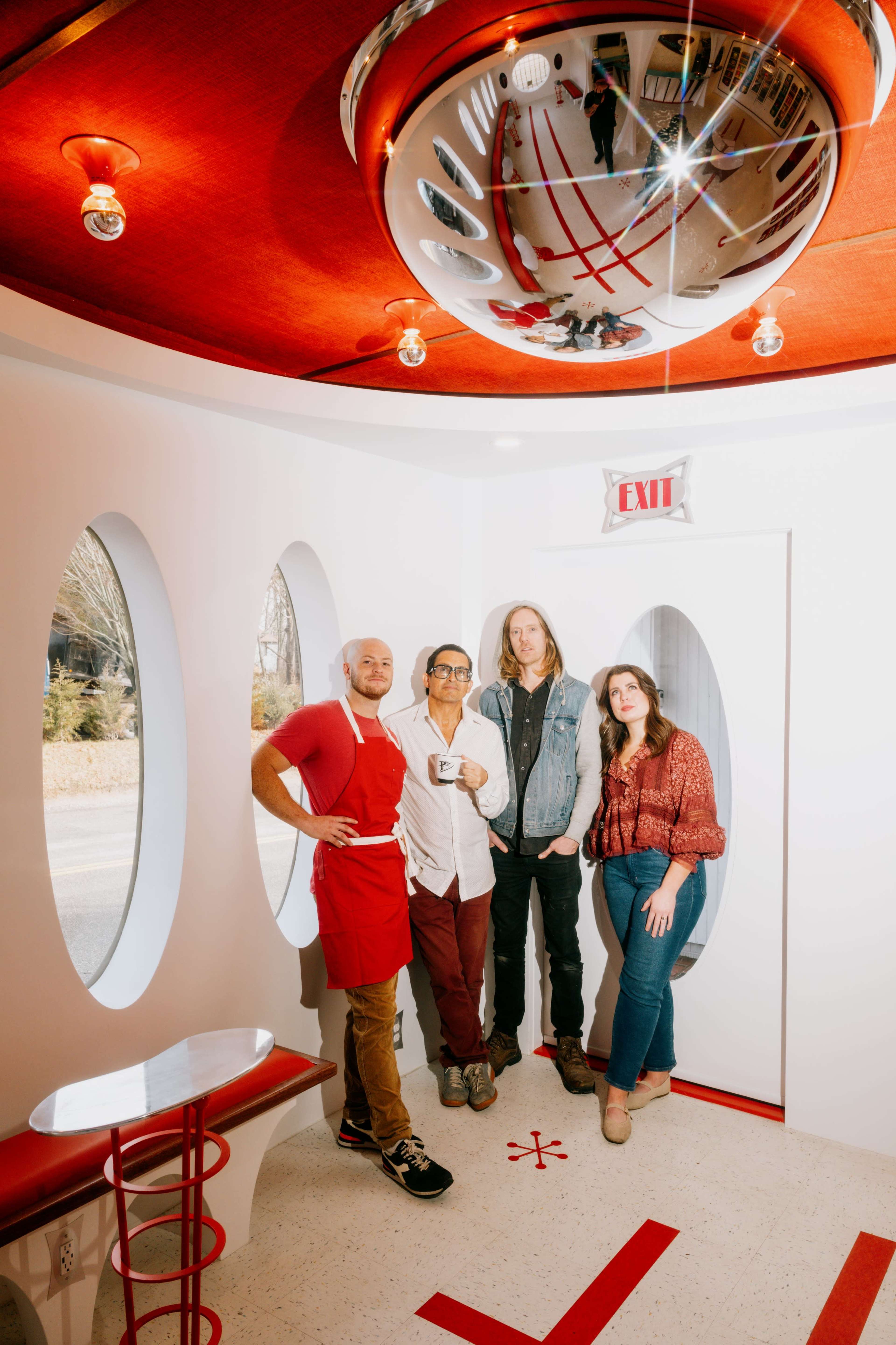 A group of four people stands inside a brightly lit, modern interior with circular windows and a large, reflective disco ball overhead.