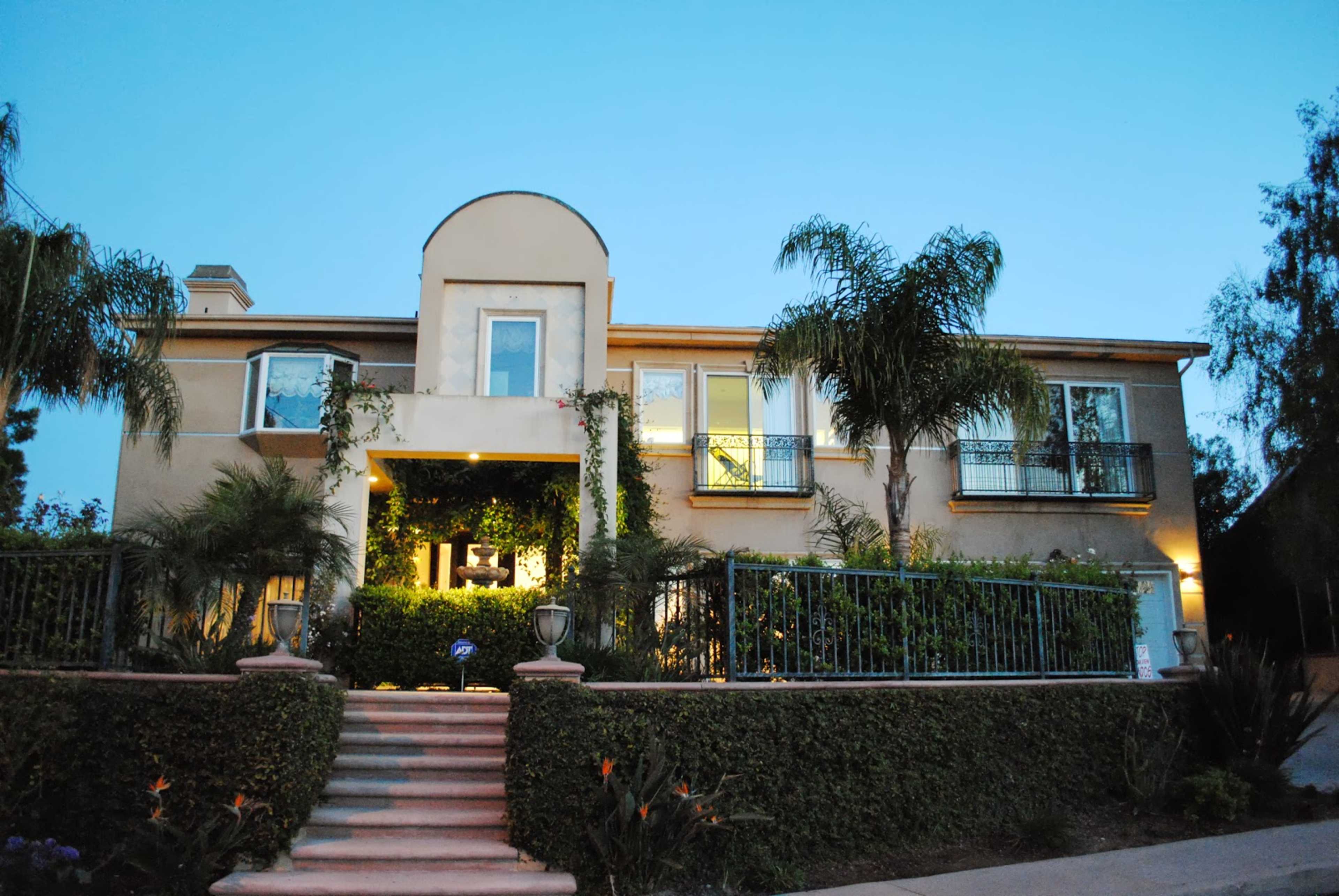 A two-story house with a Mediterranean architectural style, surrounded by landscaped greenery and palm trees, is depicted at twilight.