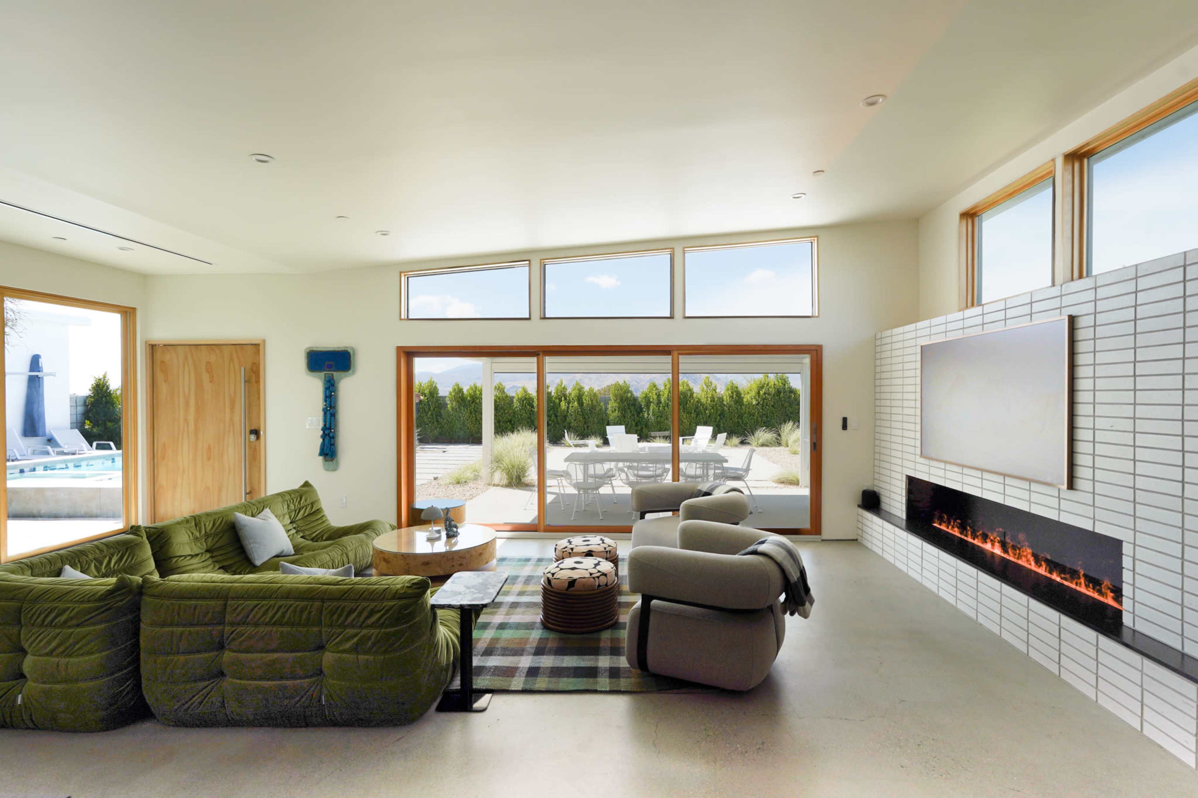 A modern living room features a green sectional sofa, a round wooden table, and large windows that overlook a patio with outdoor seating.