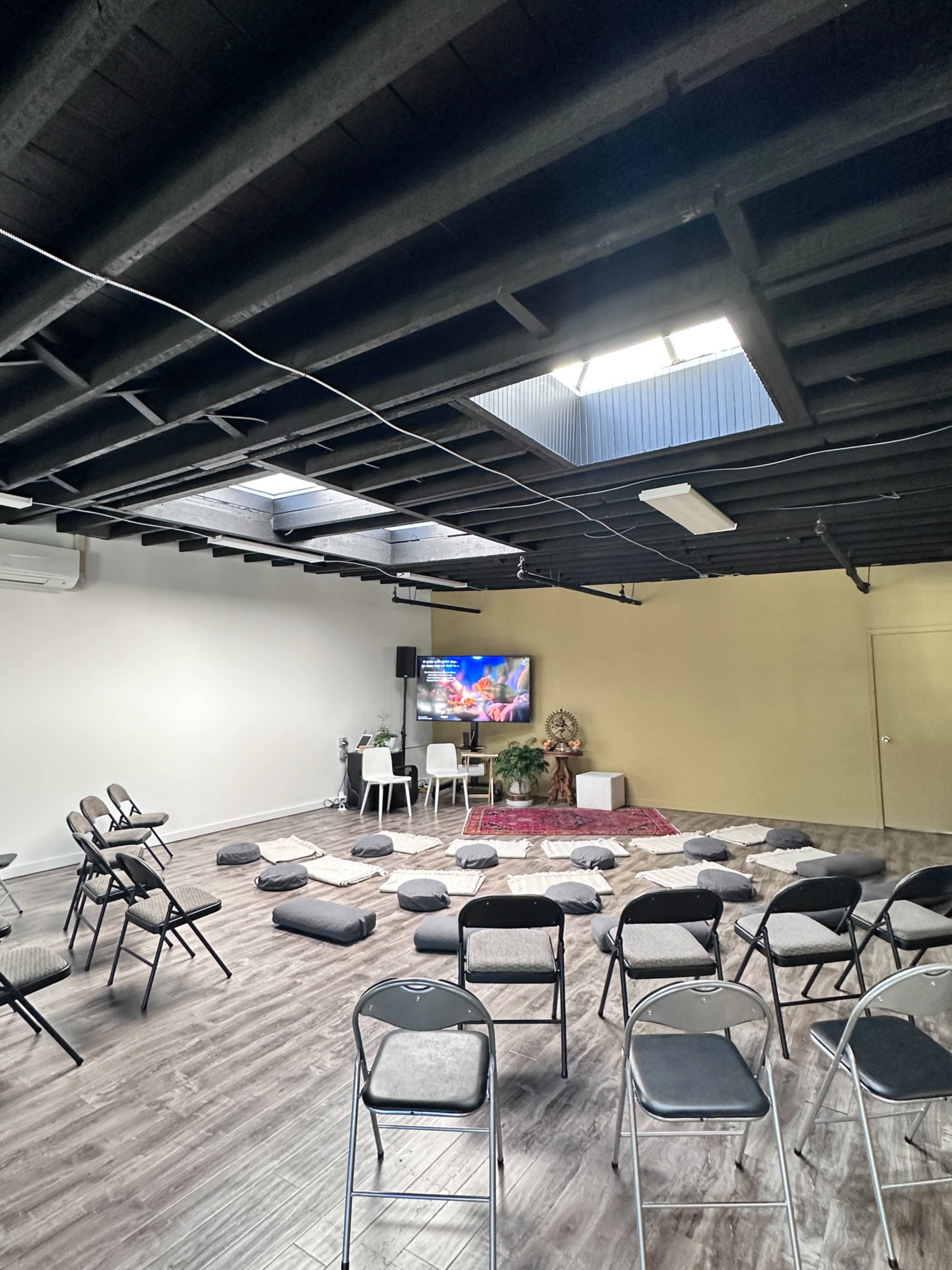 The image shows an interior space set up for a gathering, featuring chairs arranged in a semi-circle around a small stage area with cushions and a television screen.
