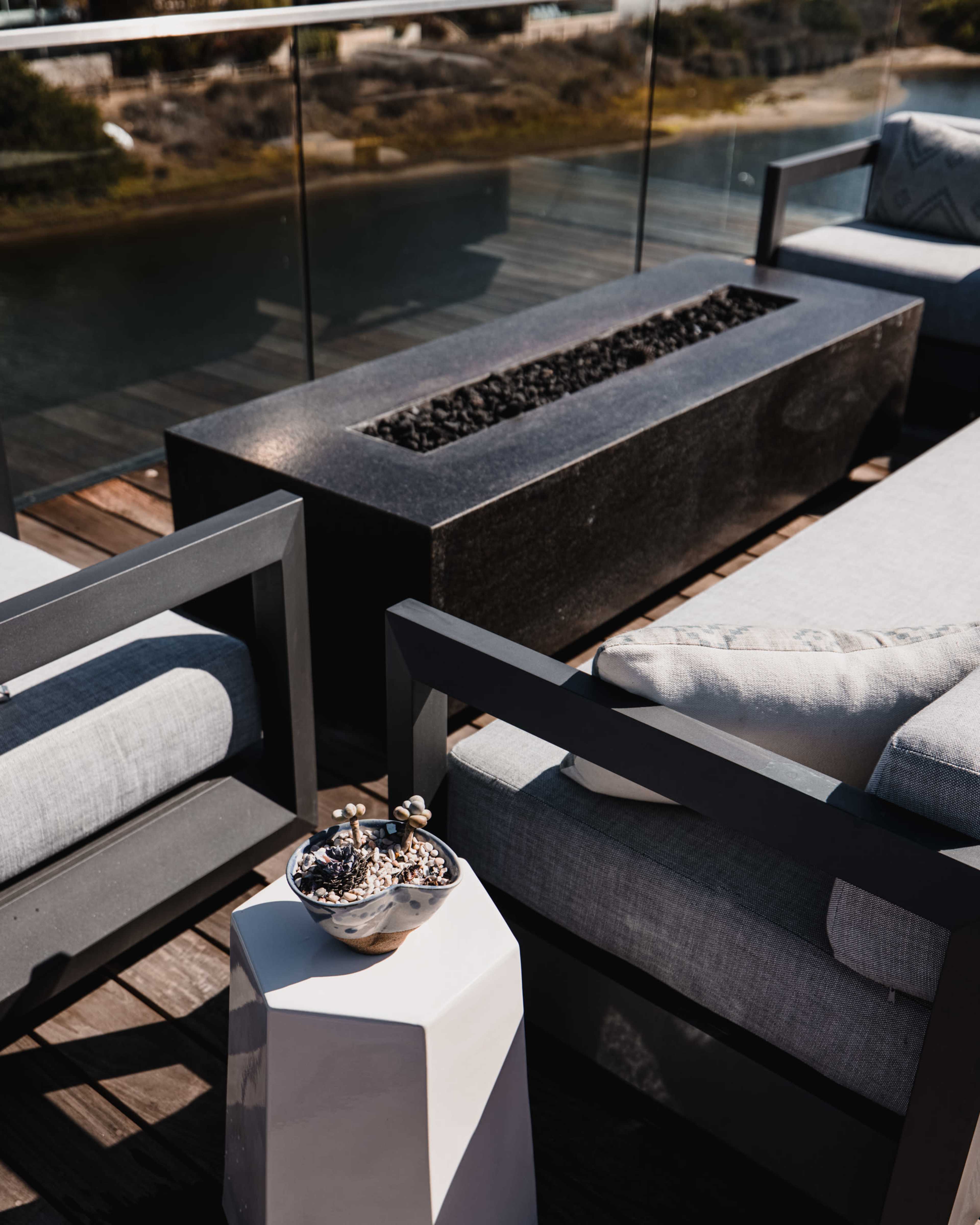 A modern outdoor lounge area features two gray chairs and a sleek fire table, with a small white table holding a bowl of snacks.