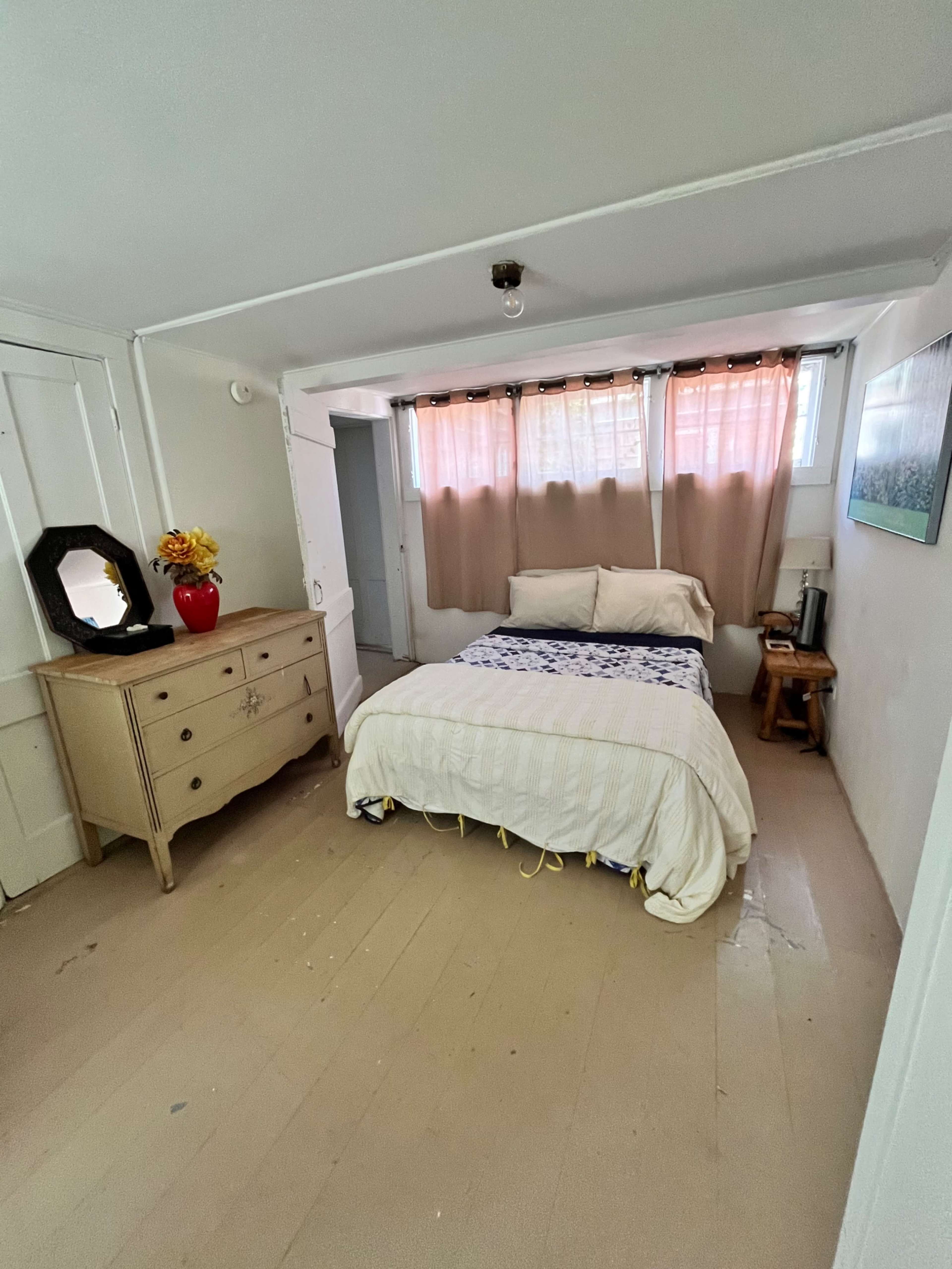 The image shows a small, simple bedroom with a bed, a wooden dresser, and natural light coming through sheer curtains.