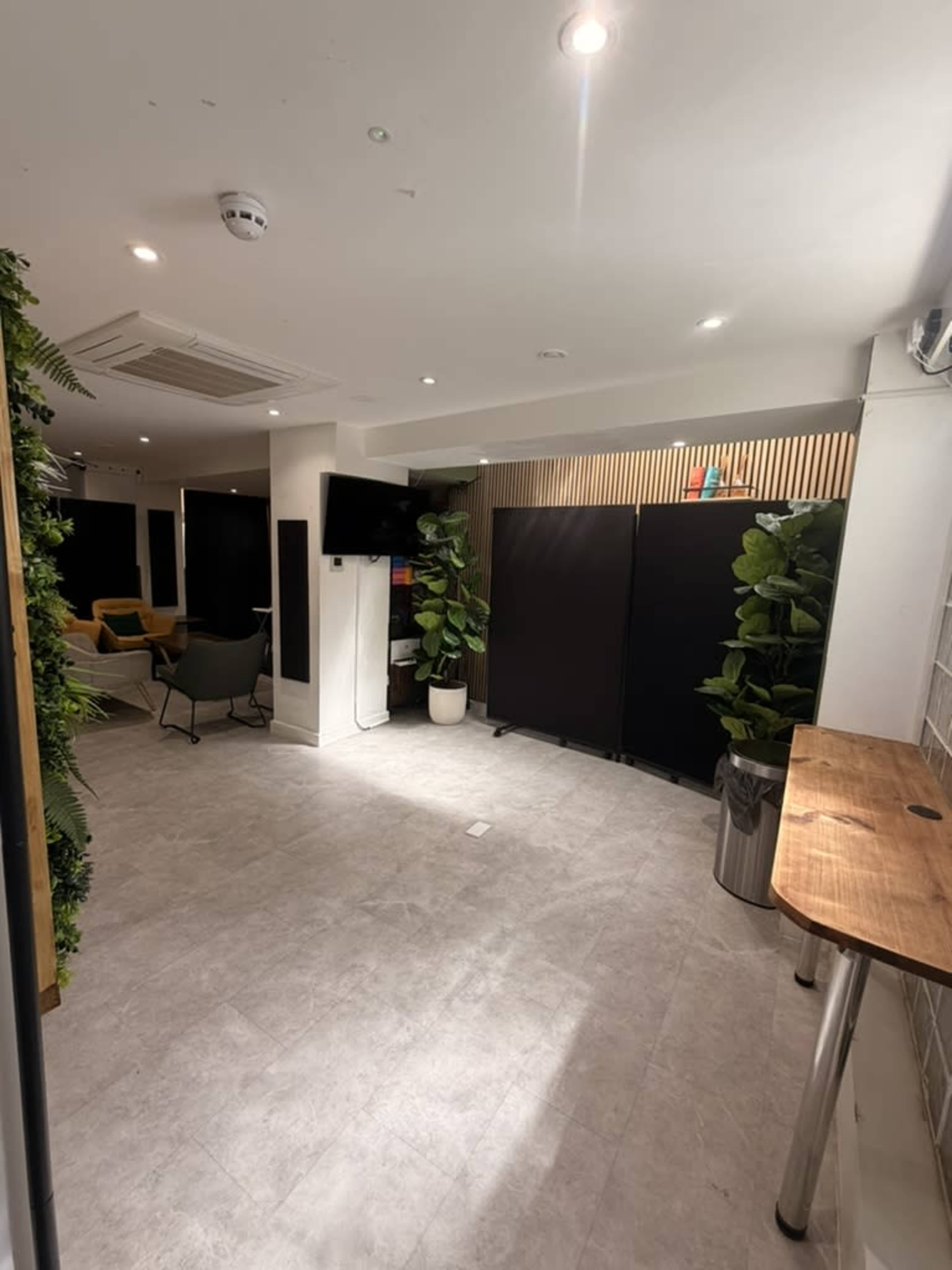 The image shows a well-lit, modern indoor space with light grey tile flooring, potted plants, a television on the wall, and a wooden counter.