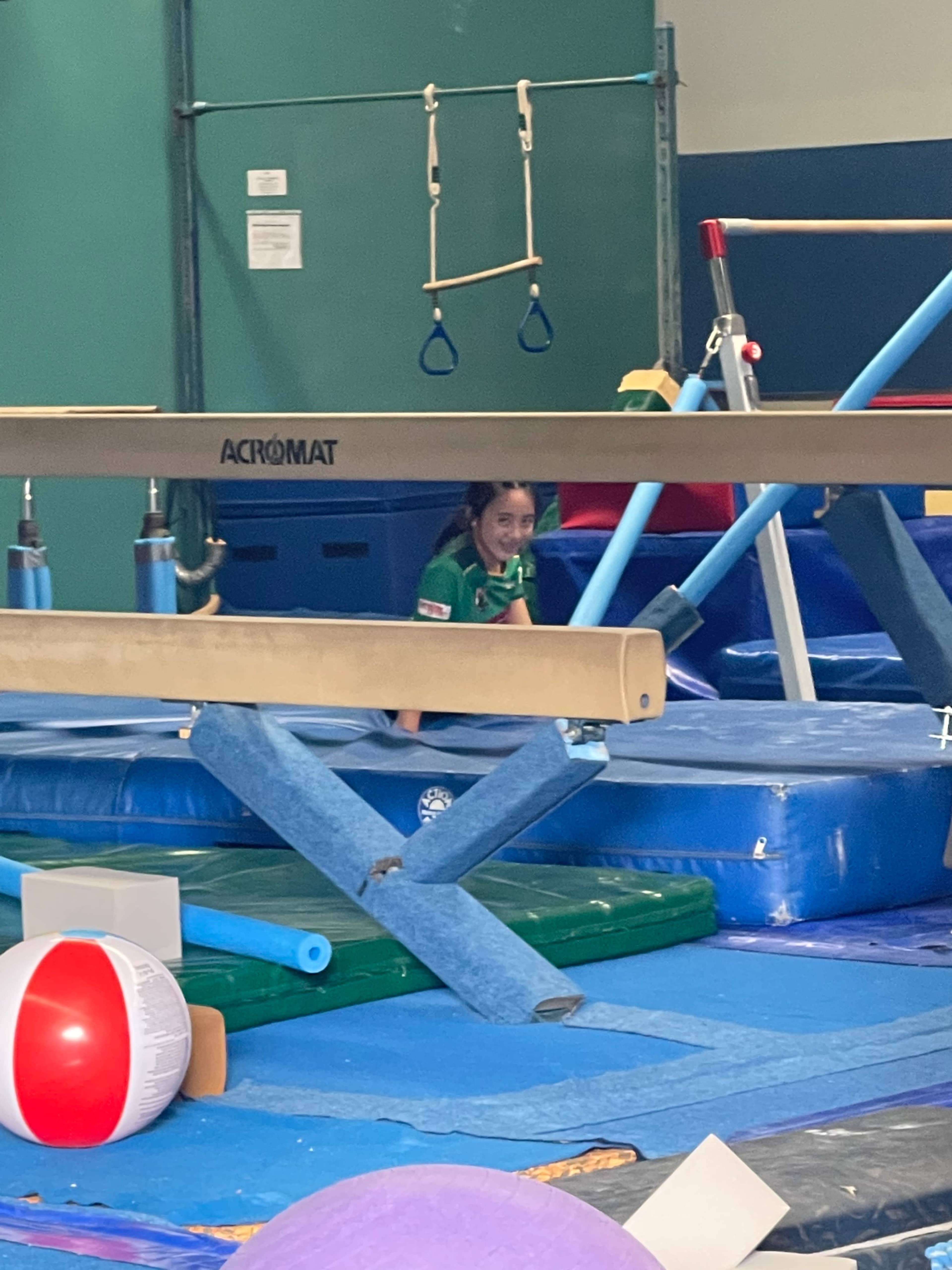 A young gymnast is practicing in a gymnasium, positioned near a balance beam and various colorful gymnastic equipment.