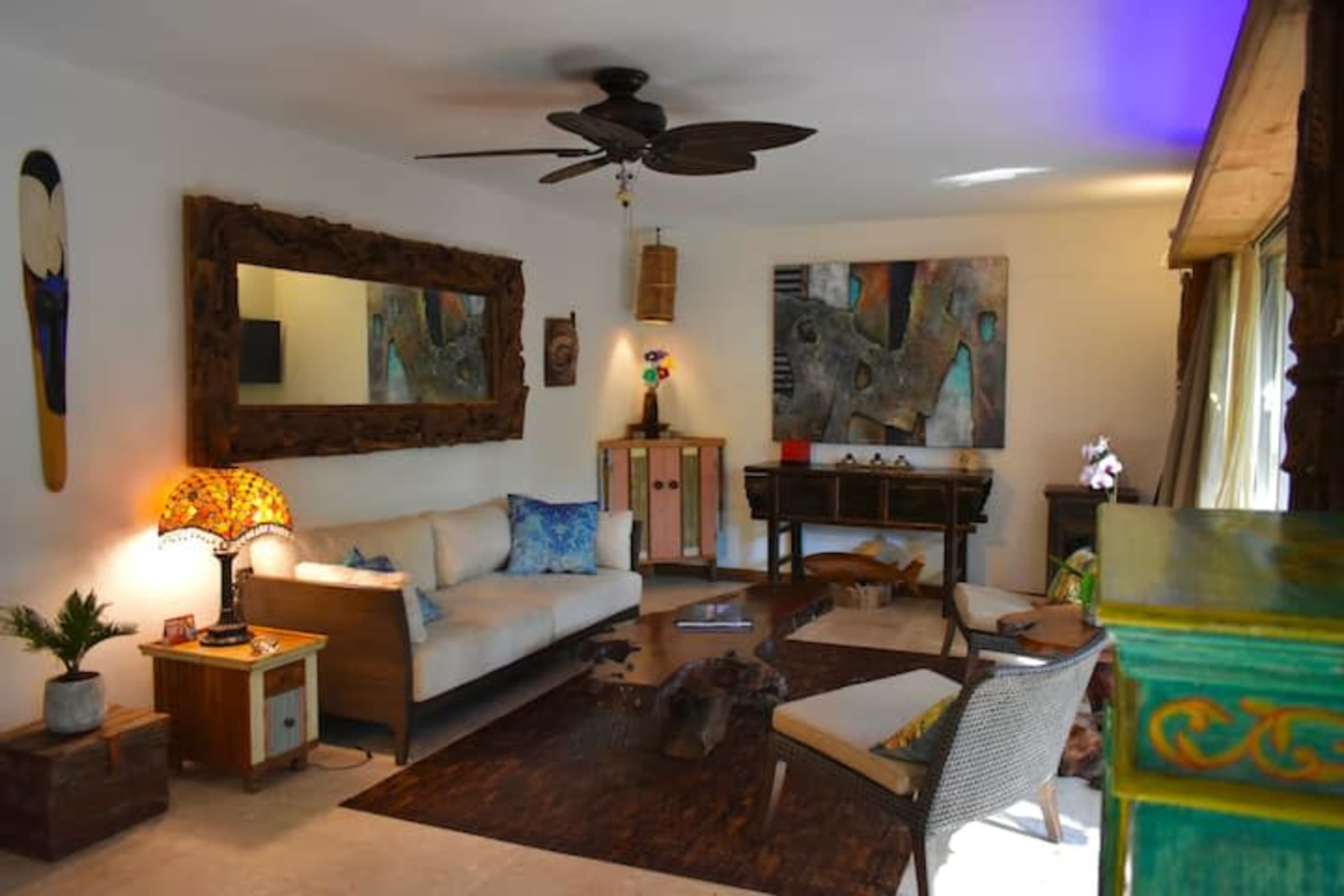 The image shows a light-filled living room featuring a beige sofa, wooden furniture, decorative art pieces, and a ceiling fan.