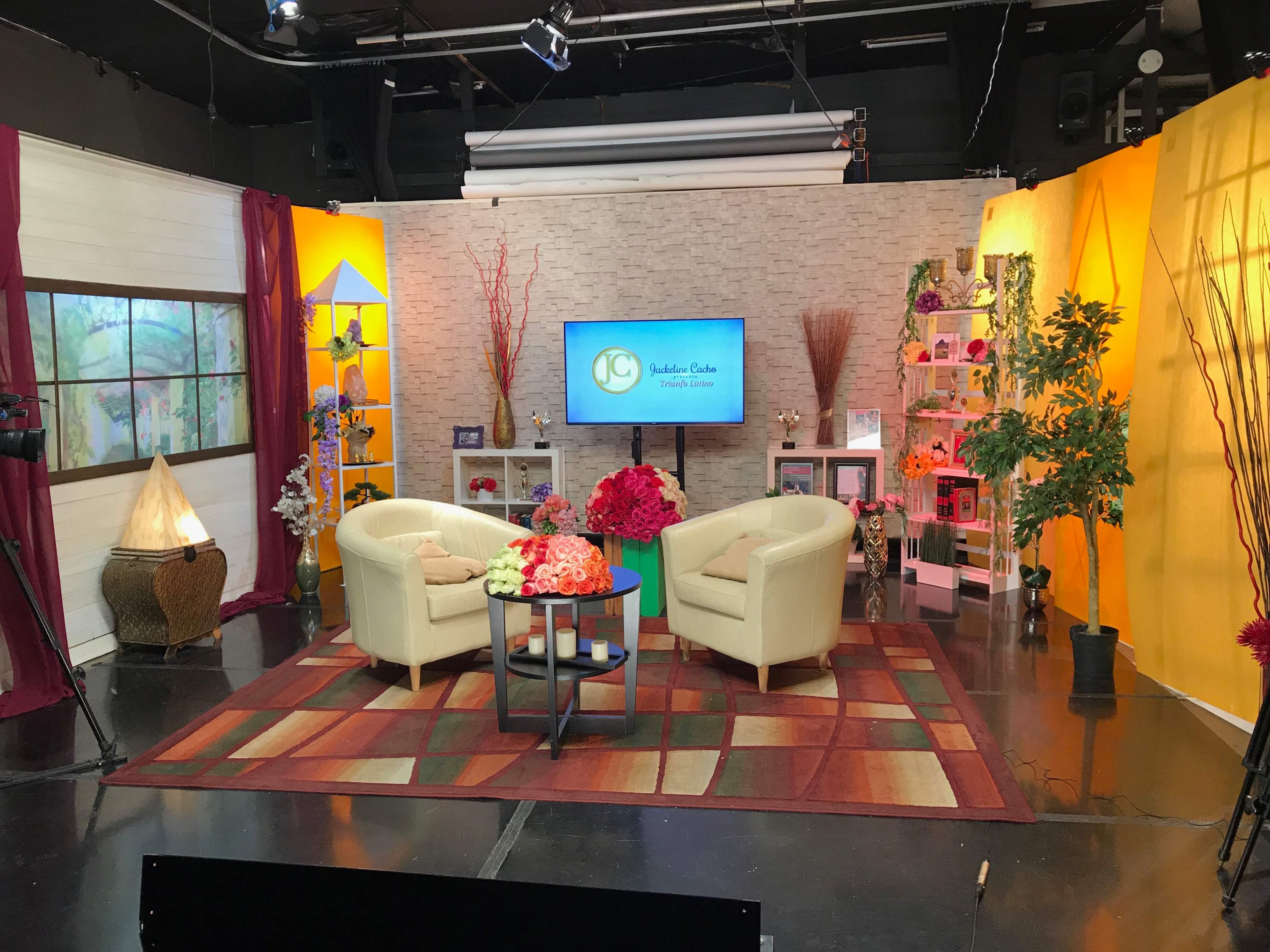 The image shows a television studio set featuring two cream-colored chairs and a round table, surrounded by colorful decorations and potted plants.