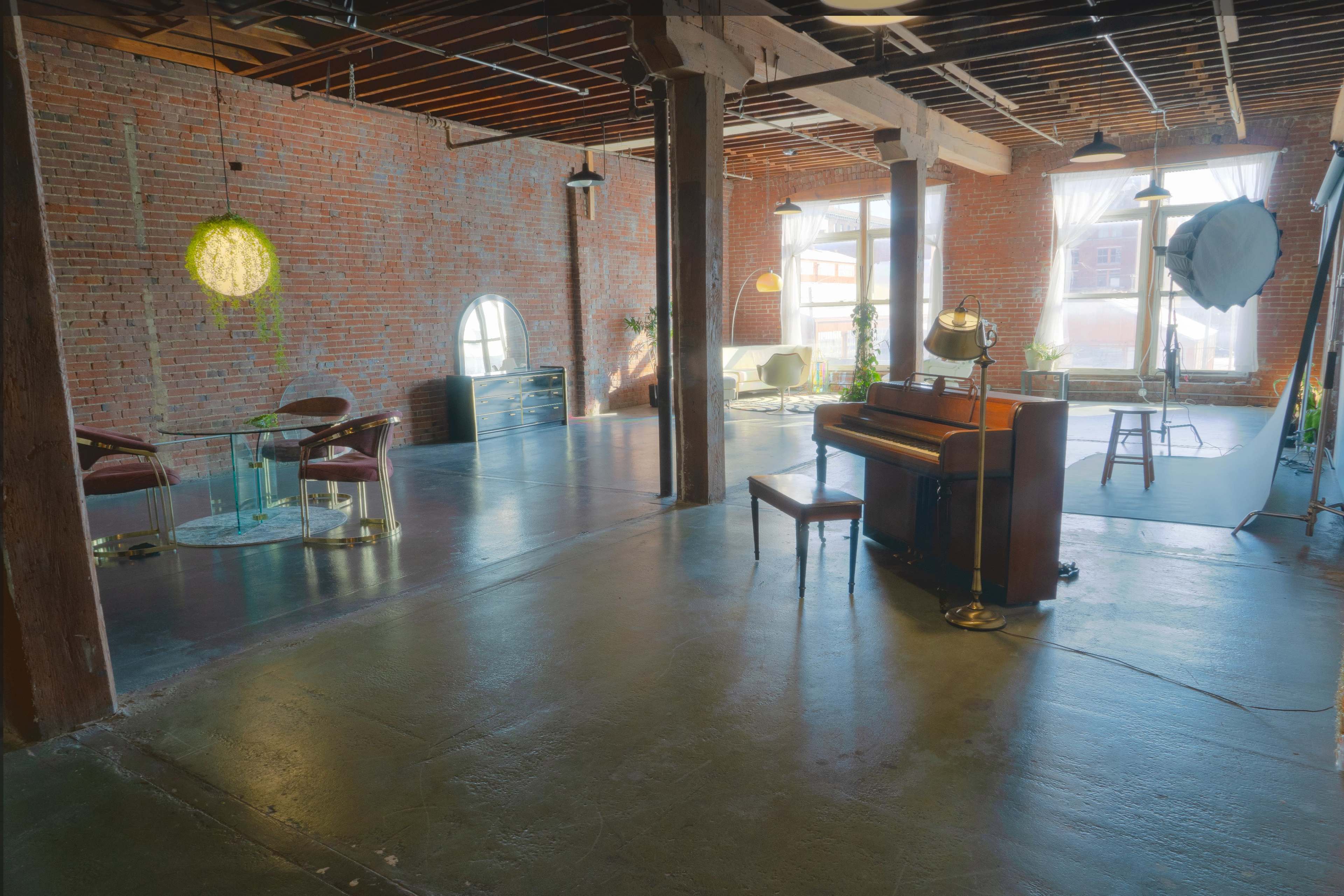 The image shows a spacious industrial-style loft with exposed brick walls, wooden beams, and a mix of modern furniture, including a piano and a glass dining table.