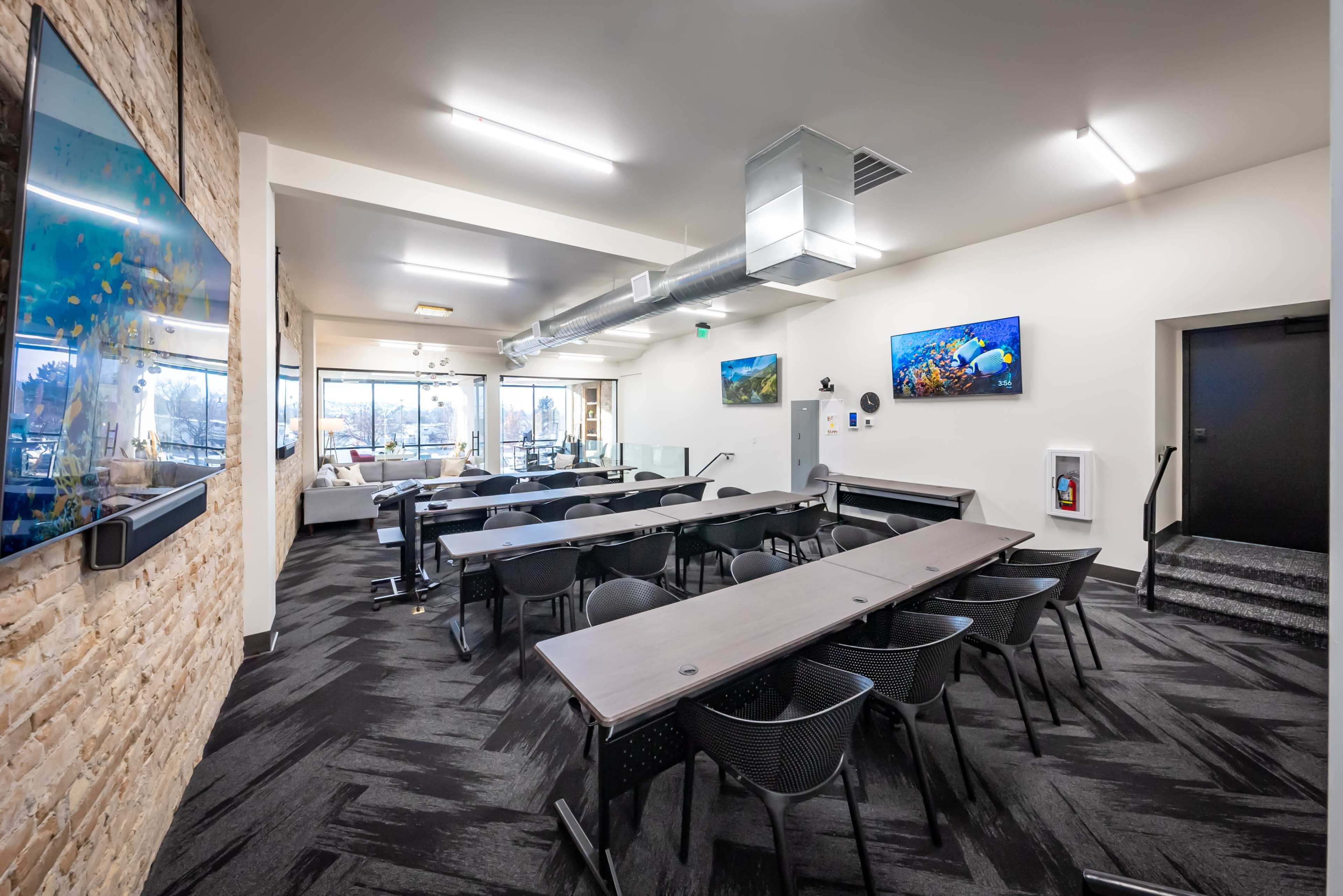 A modern conference room with several tables and chairs arranged for meetings, featuring large windows and wall-mounted screens displaying underwater scenes.