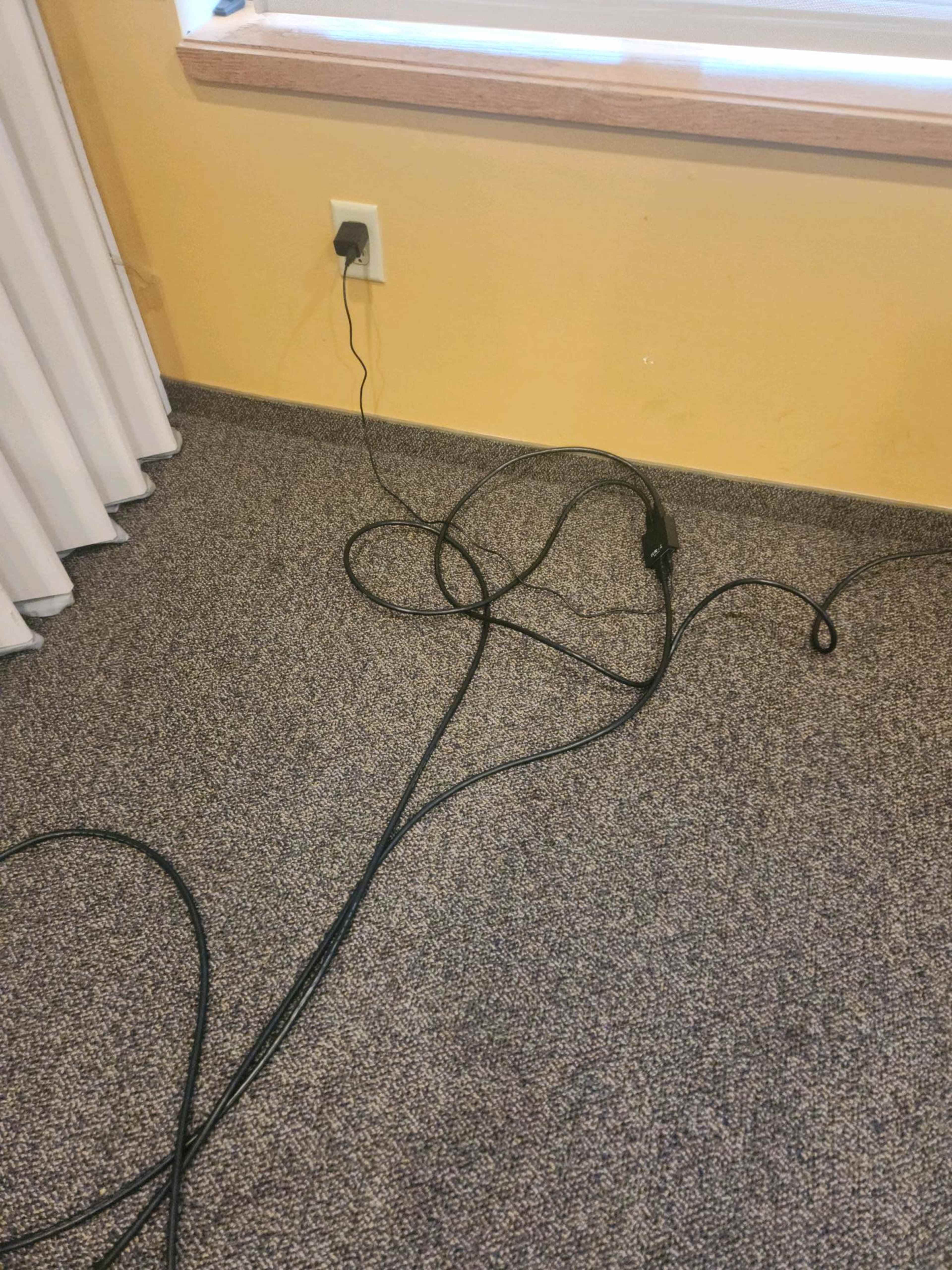 A tangle of black electrical cables runs across a carpeted floor near a wall plug.