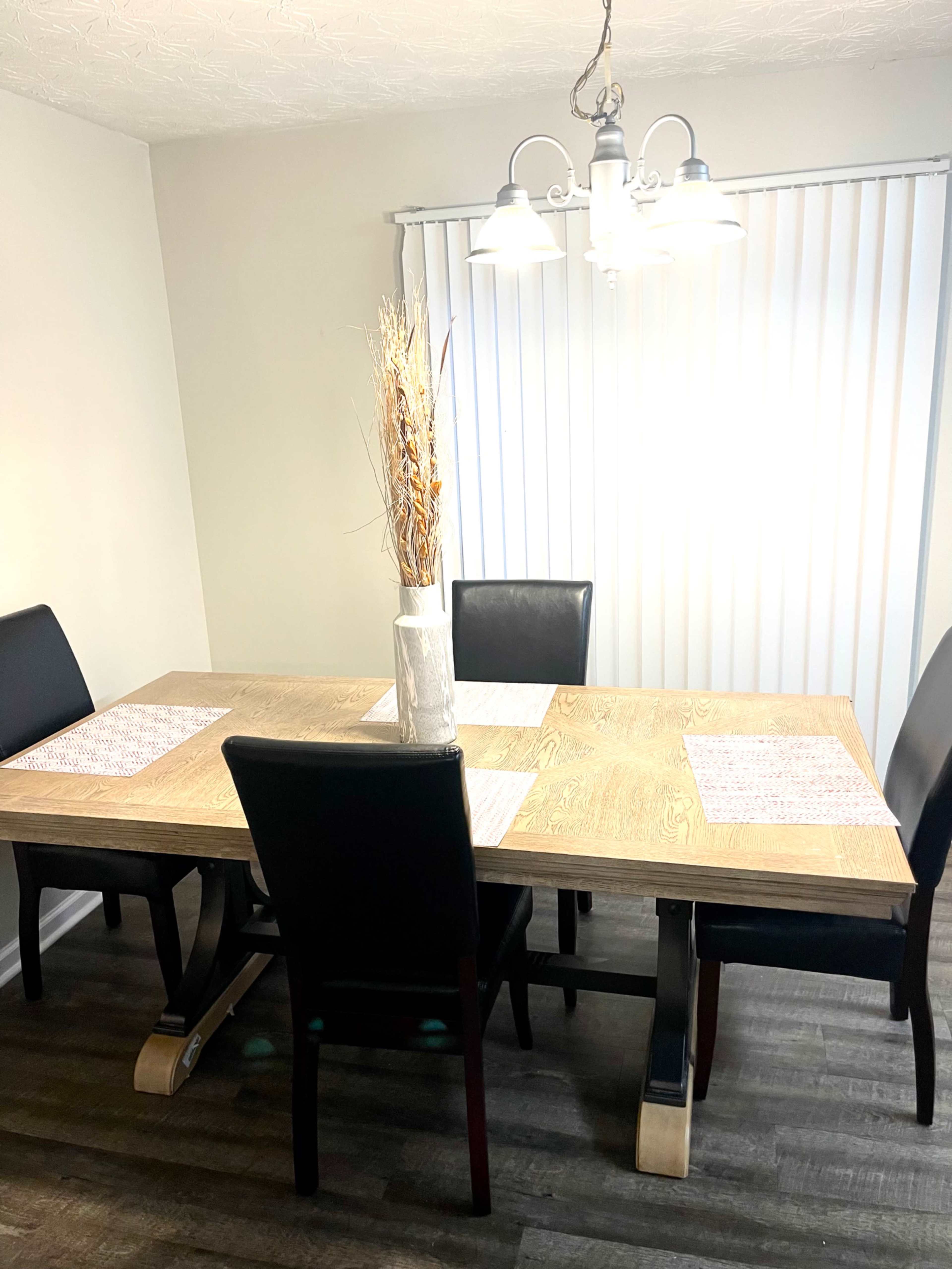 A dining table with four black chairs is set in a room featuring light-colored walls and vertical blinds.