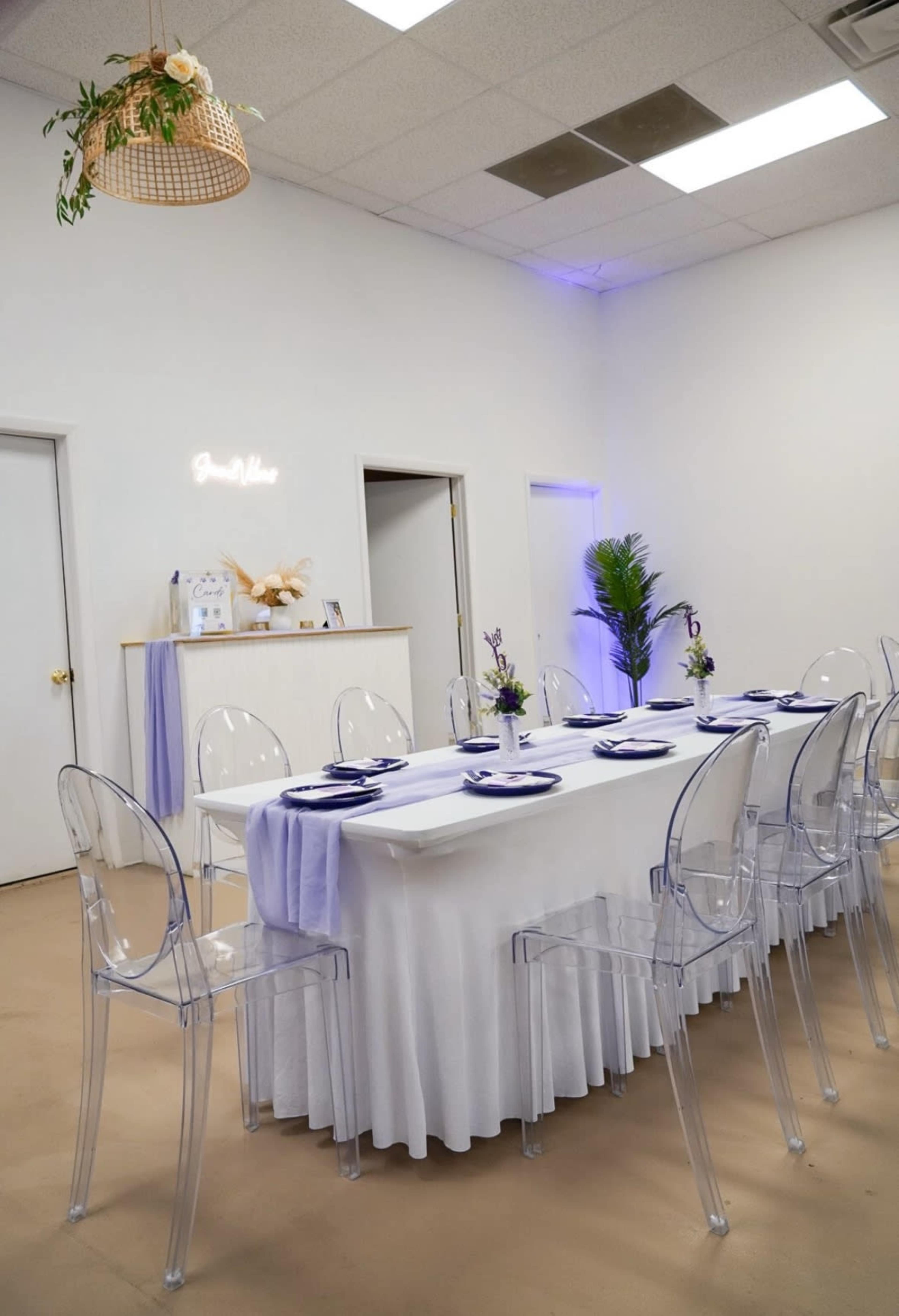 A long dining table is set up in a brightly lit room, surrounded by clear acrylic chairs and adorned with purple table linens and place settings.