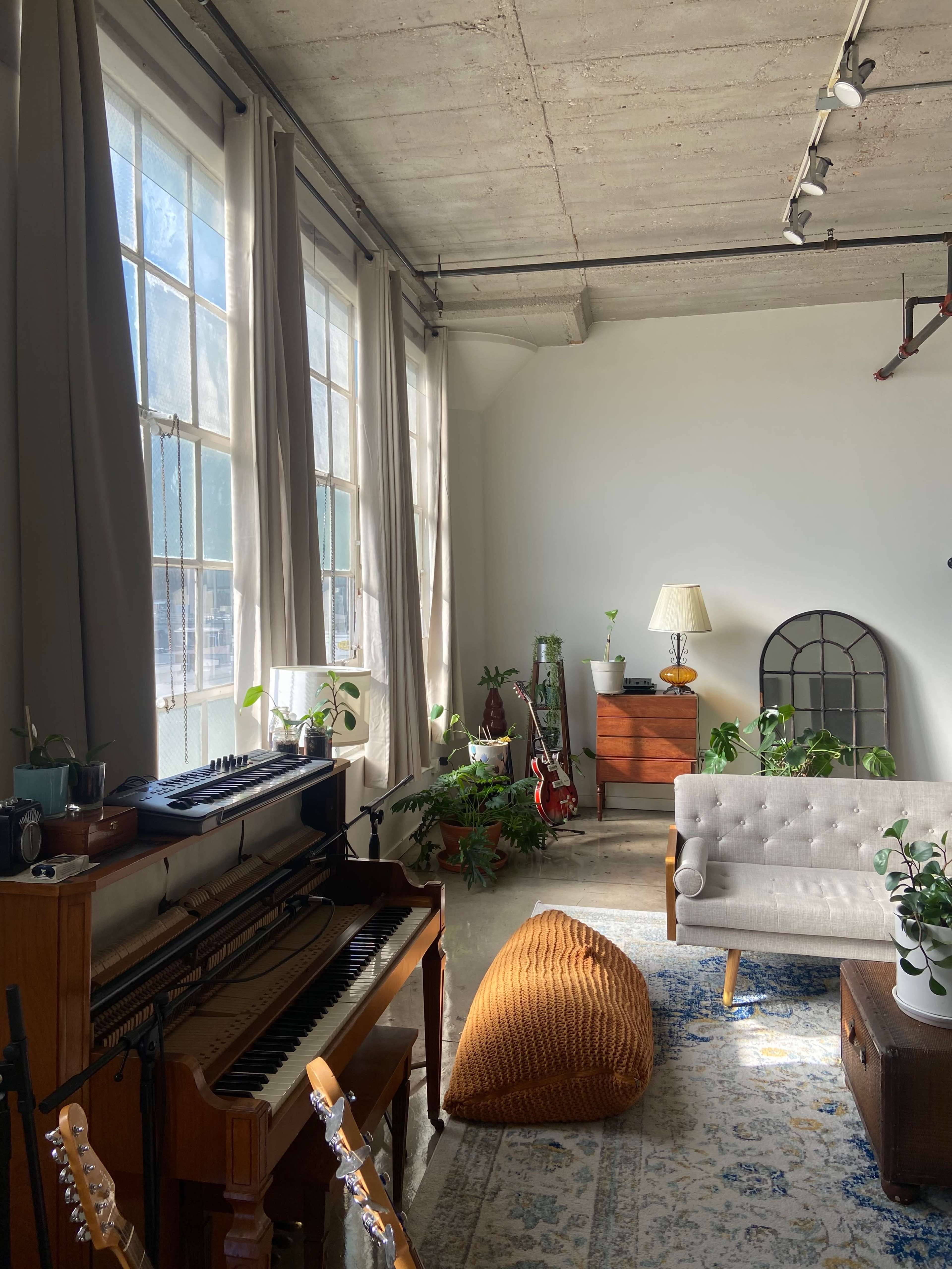 The image shows a bright room with large windows, featuring a piano, a grey sofa, a wooden cabinet, and various plants.
