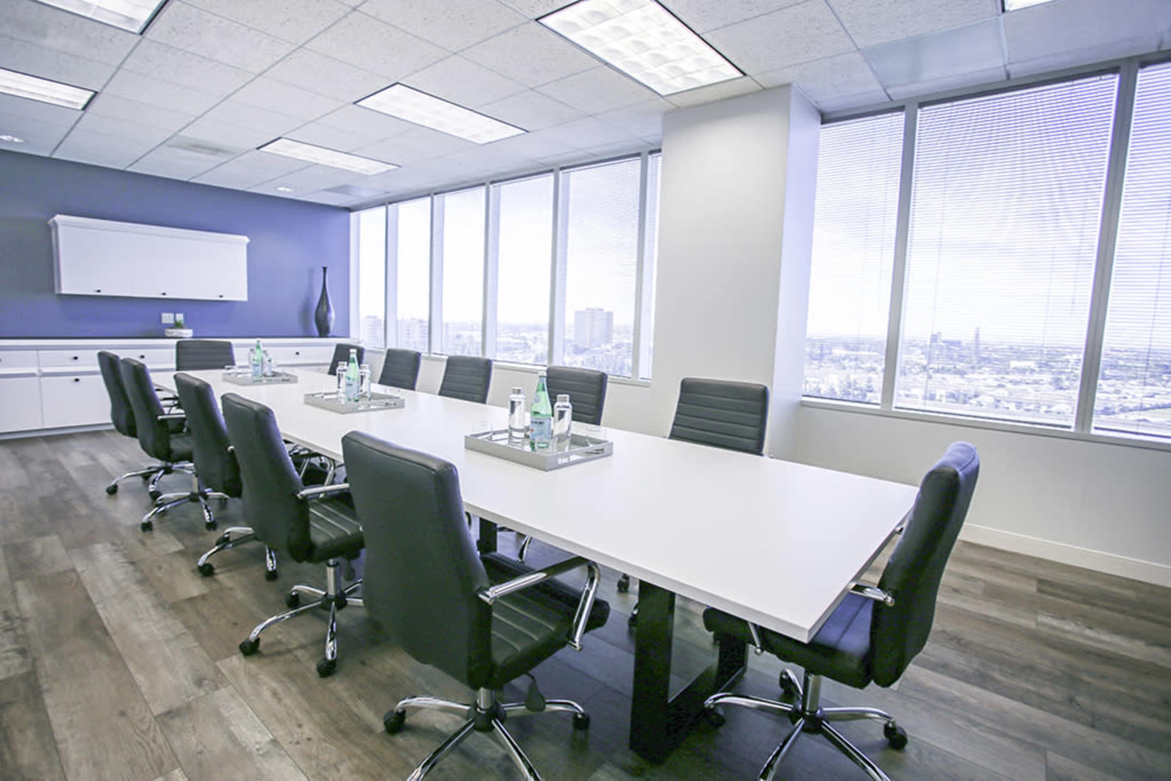 A large, modern conference room features a long white table surrounded by black chairs, with tall windows offering a view of the city skyline.