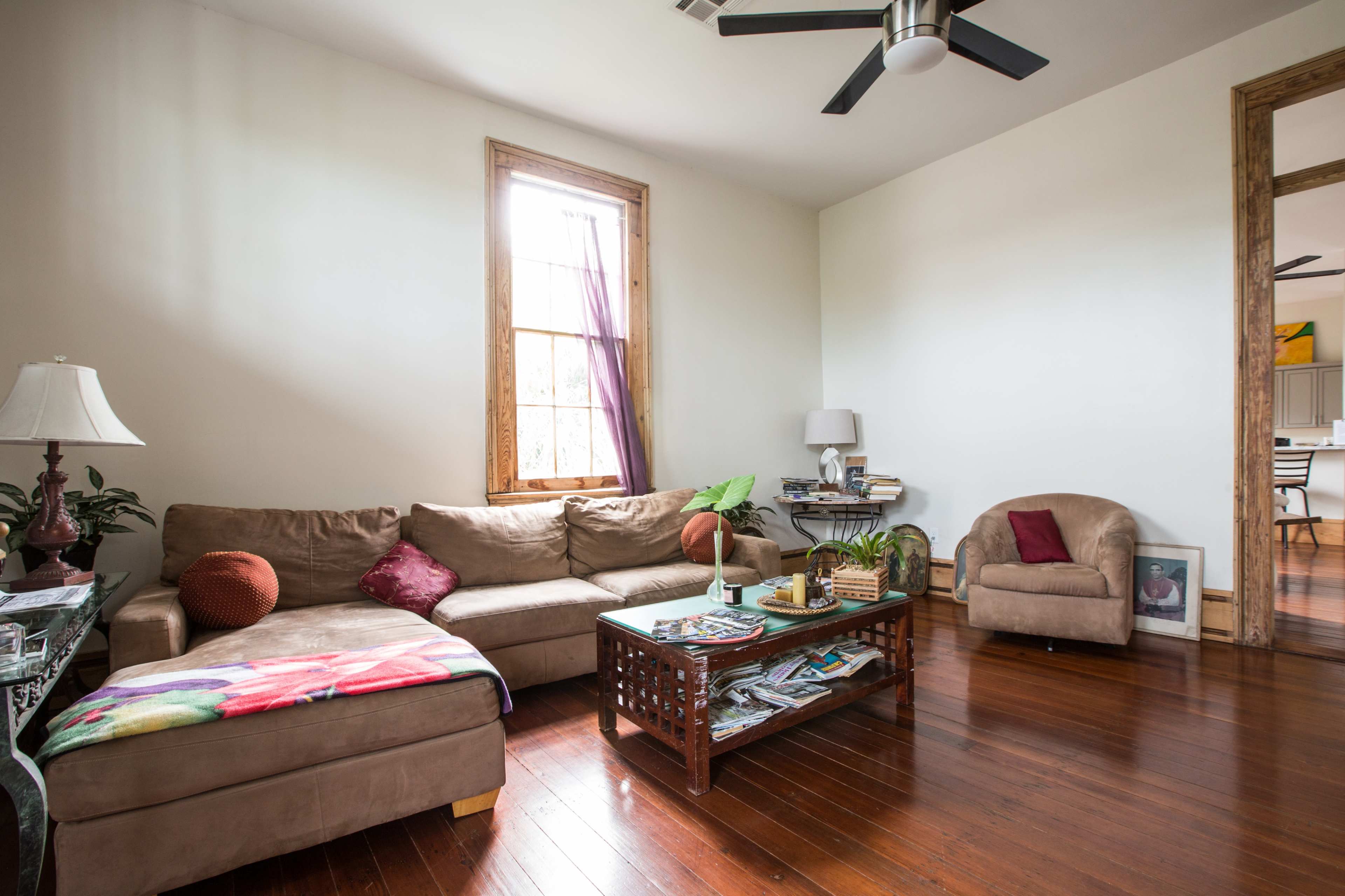 A living room features a brown sectional sofa, a small brown armchair, a wooden coffee table with magazines on it, and a window with purple curtains.