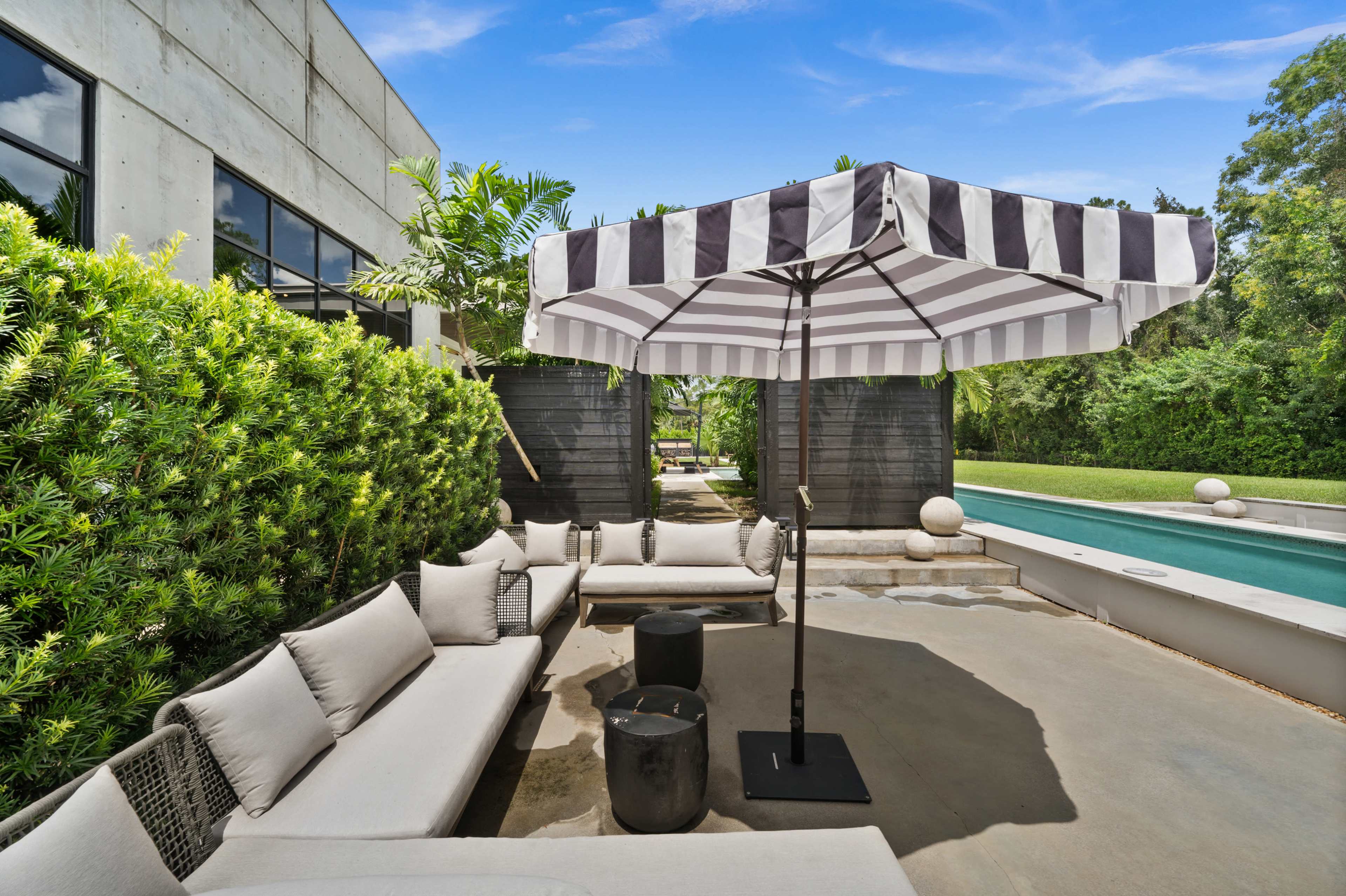 A shaded seating area with a striped umbrella overlooks a rectangular swimming pool surrounded by lush greenery.