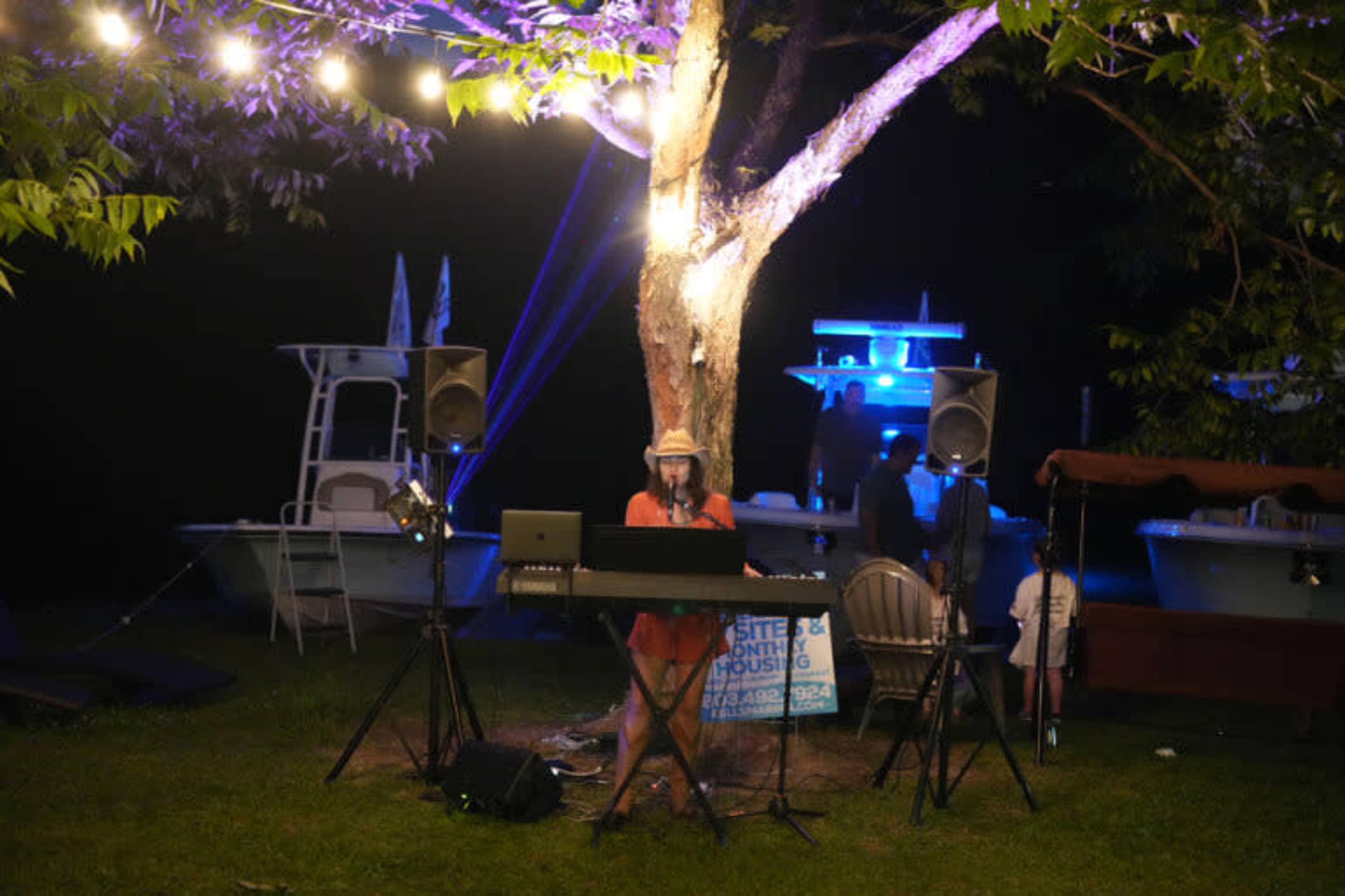A woman plays keyboard music under a tree adorned with string lights, while people gather around nearby boats and seating.