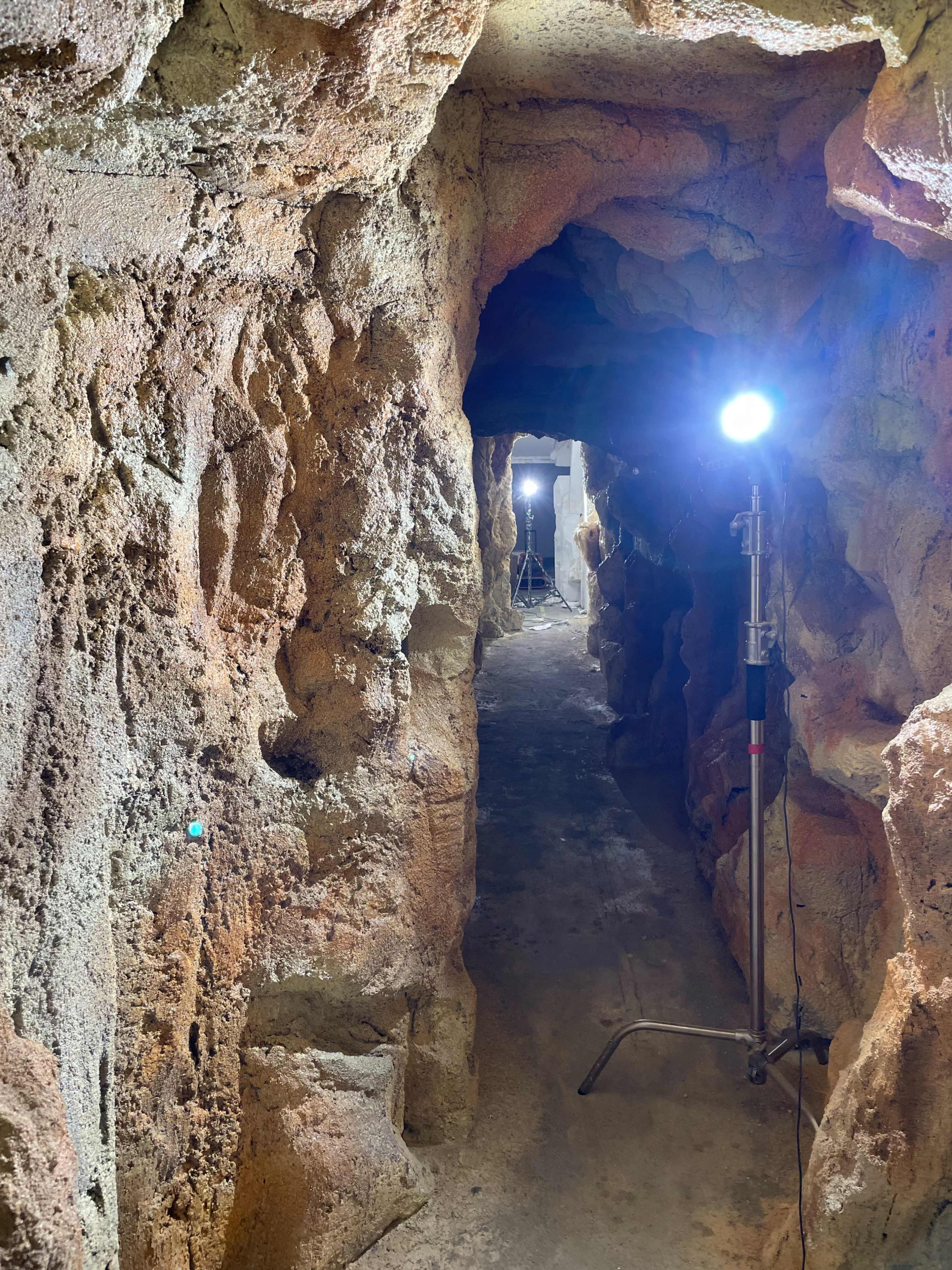 A dimly lit cave-like tunnel with rough, rocky walls and light stands positioned along the pathway.