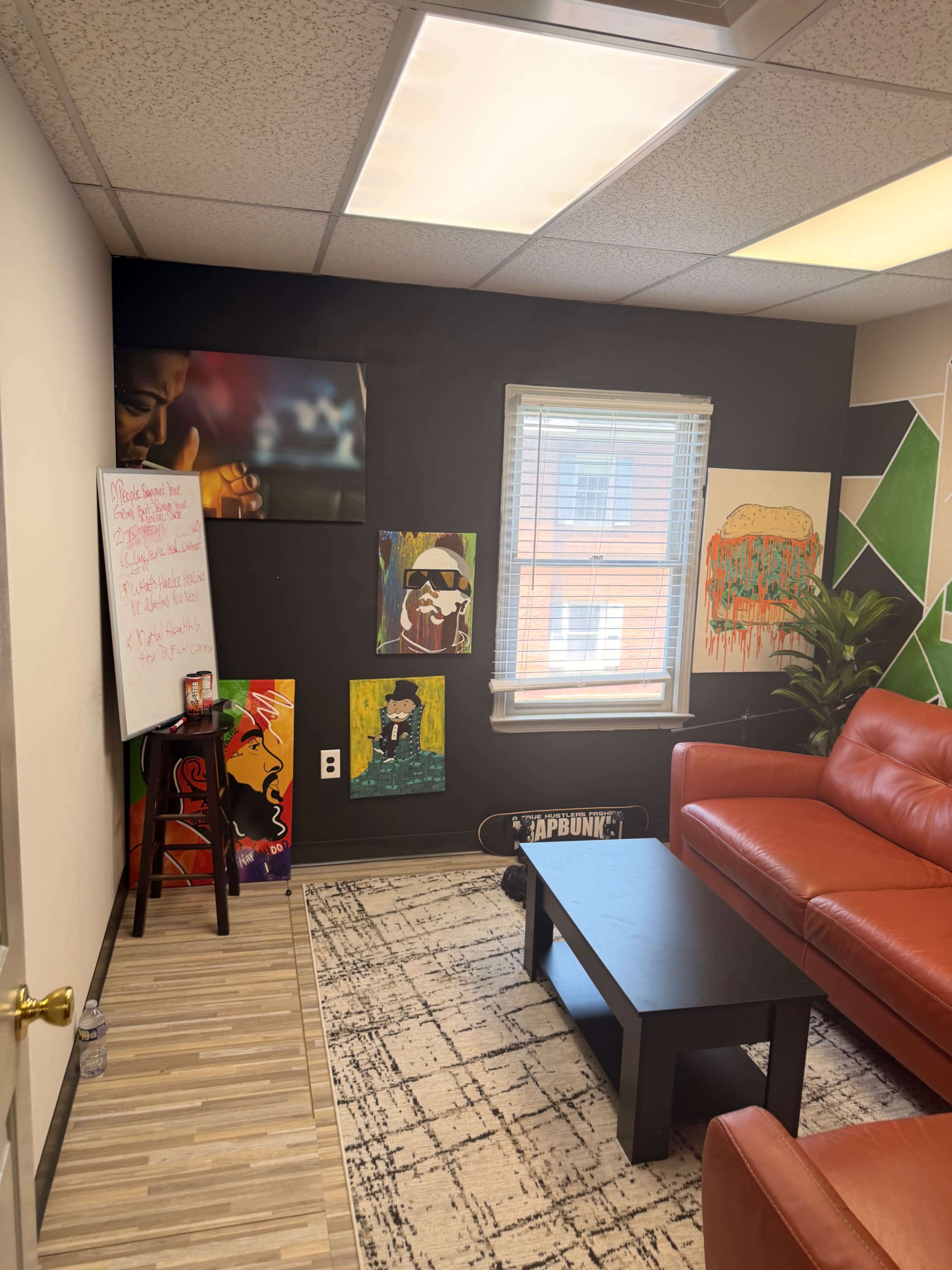 The image shows a small lounge area with a red sofa, a dark coffee table, colorful wall art, and a whiteboard beside a window.