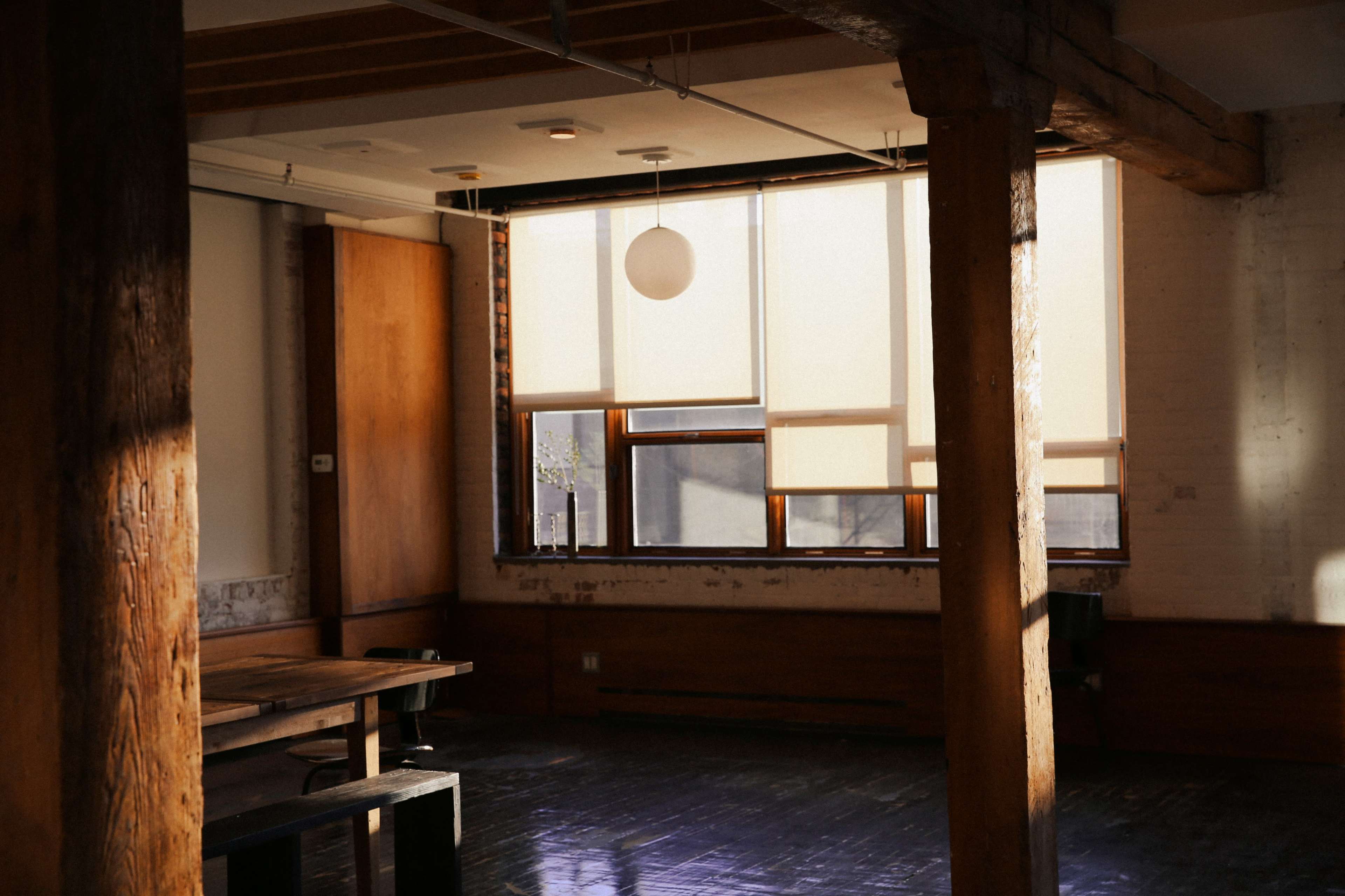 A sunlit interior features wooden beams, a large window with soft shades, and a simple wooden table.