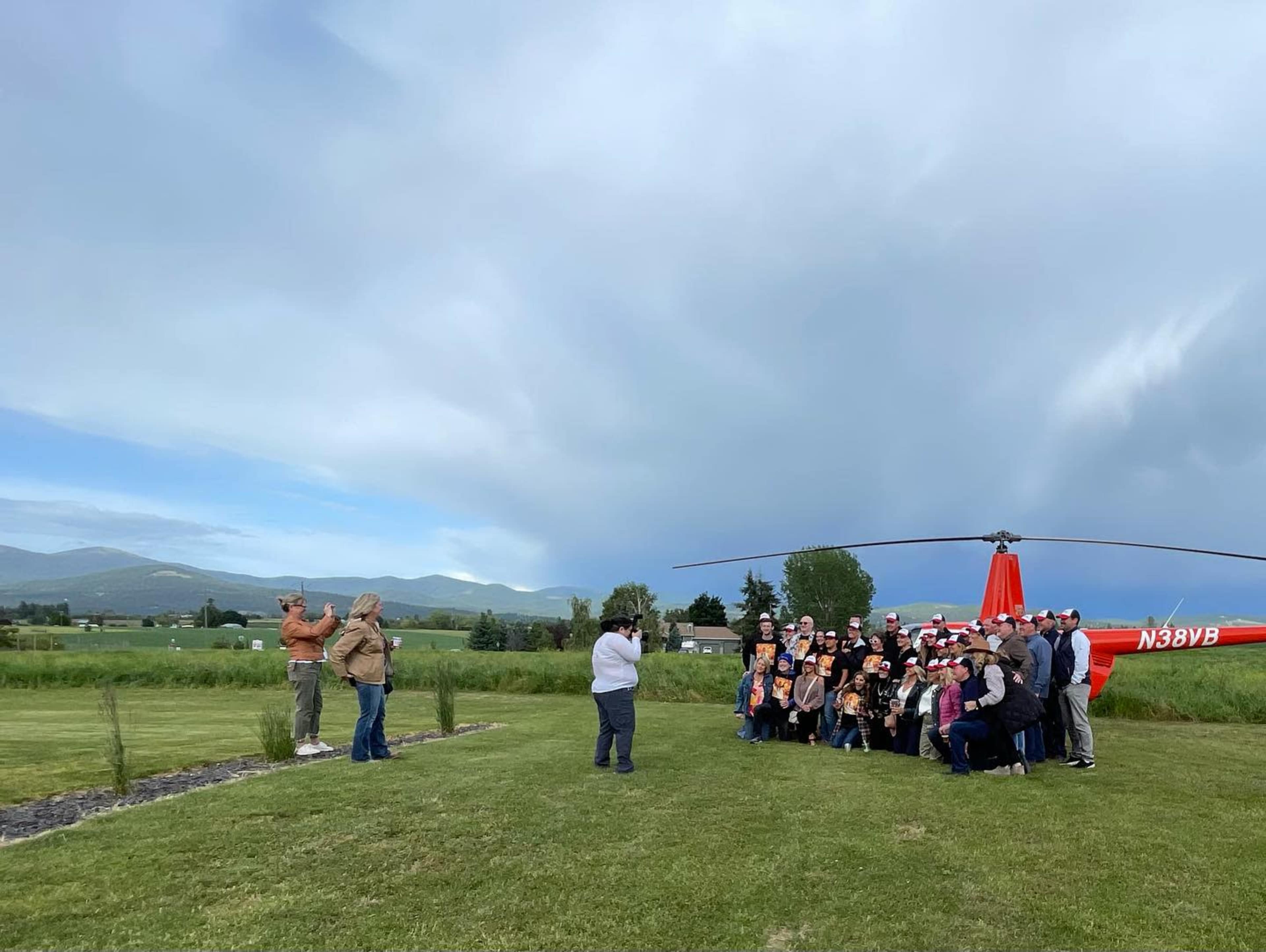 A large group of people poses for a photo in front of a red helicopter on a grassy field with mountains in the background.