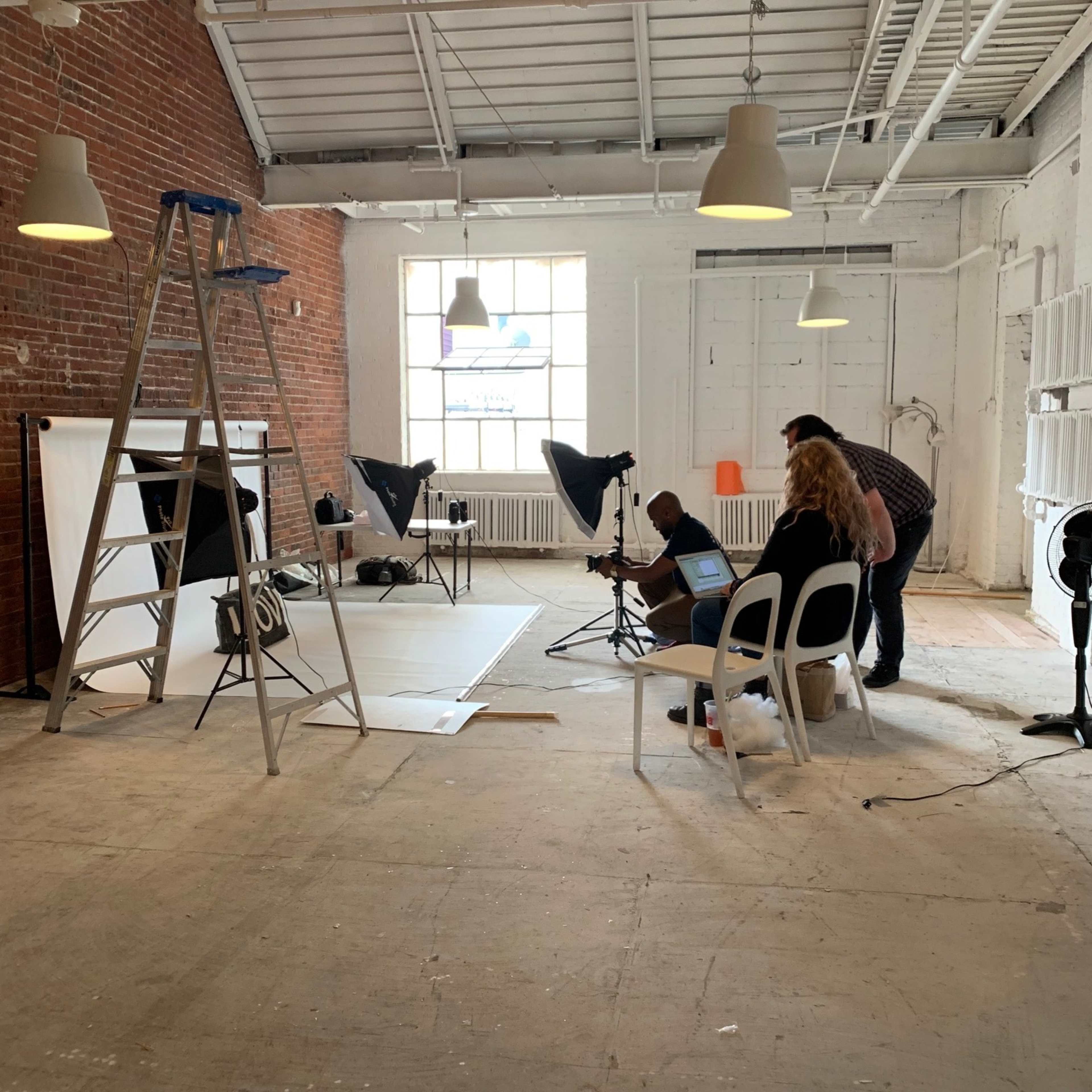 A photography studio features a ladder, lights set up on the floor, and three people preparing for a photoshoot.