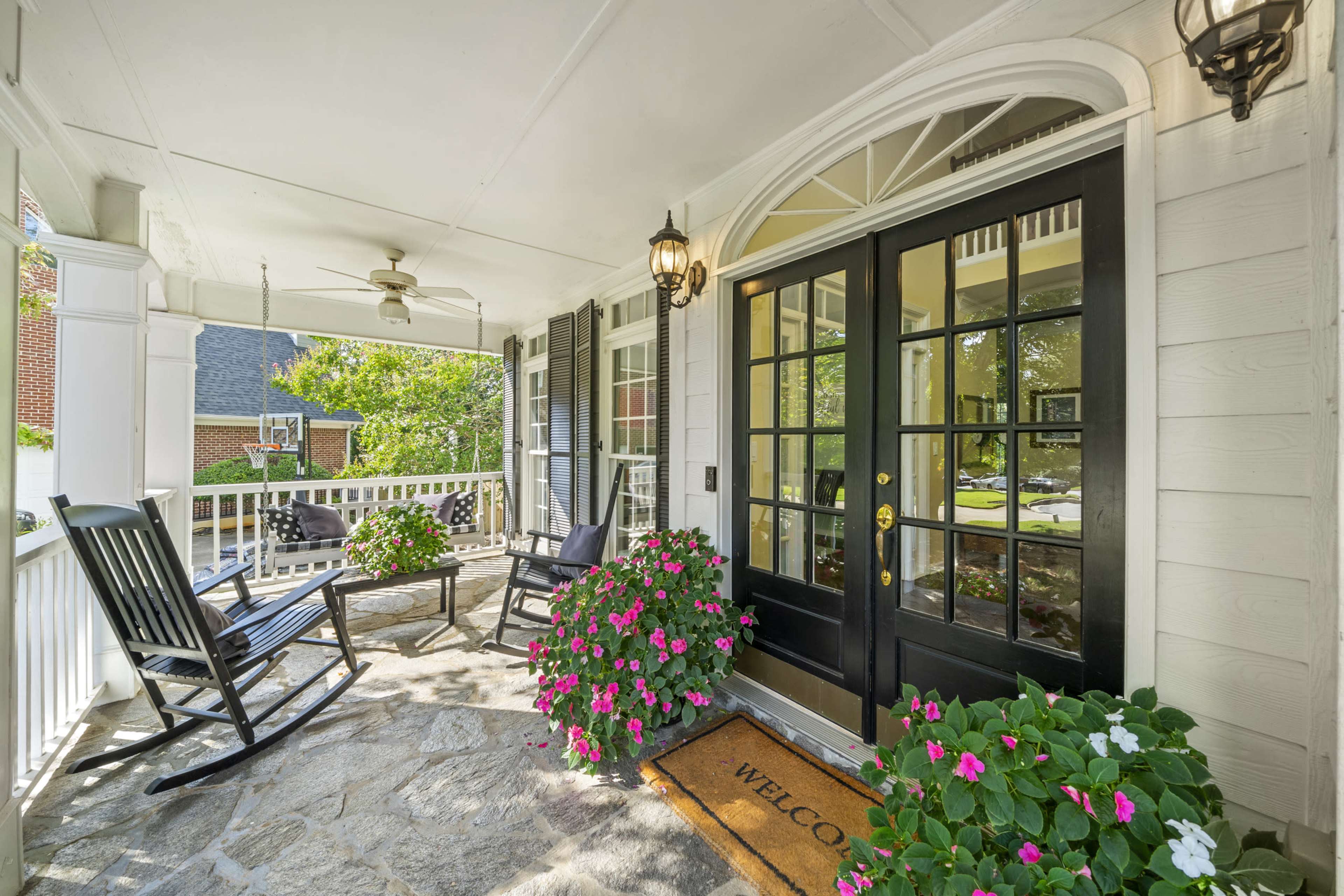 A porch with a black front door, rocking chairs, potted flowers, and a welcome mat.
