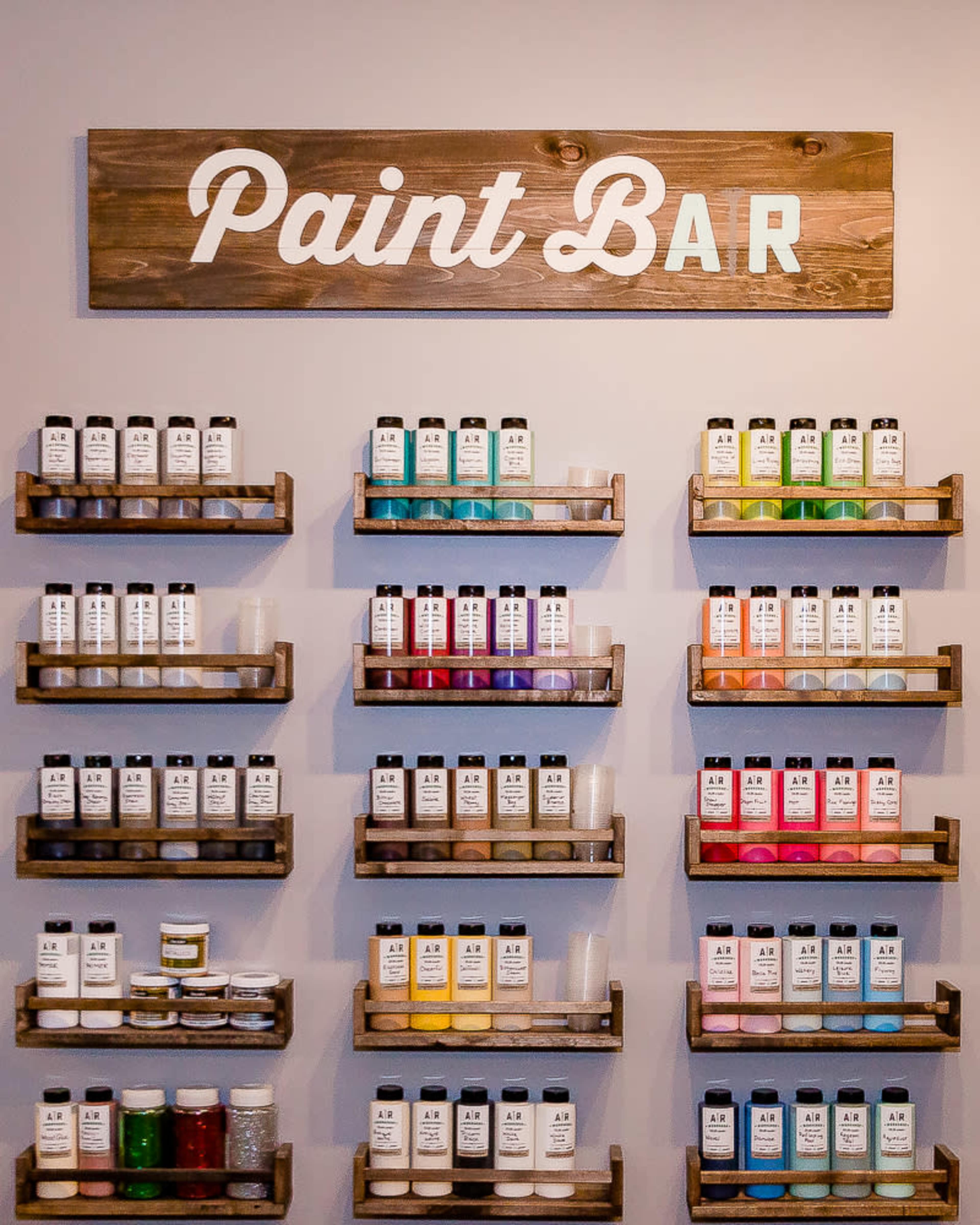 A wall display of neatly arranged paint bottles on wooden shelves, with a sign reading "Paint Bar" above them.