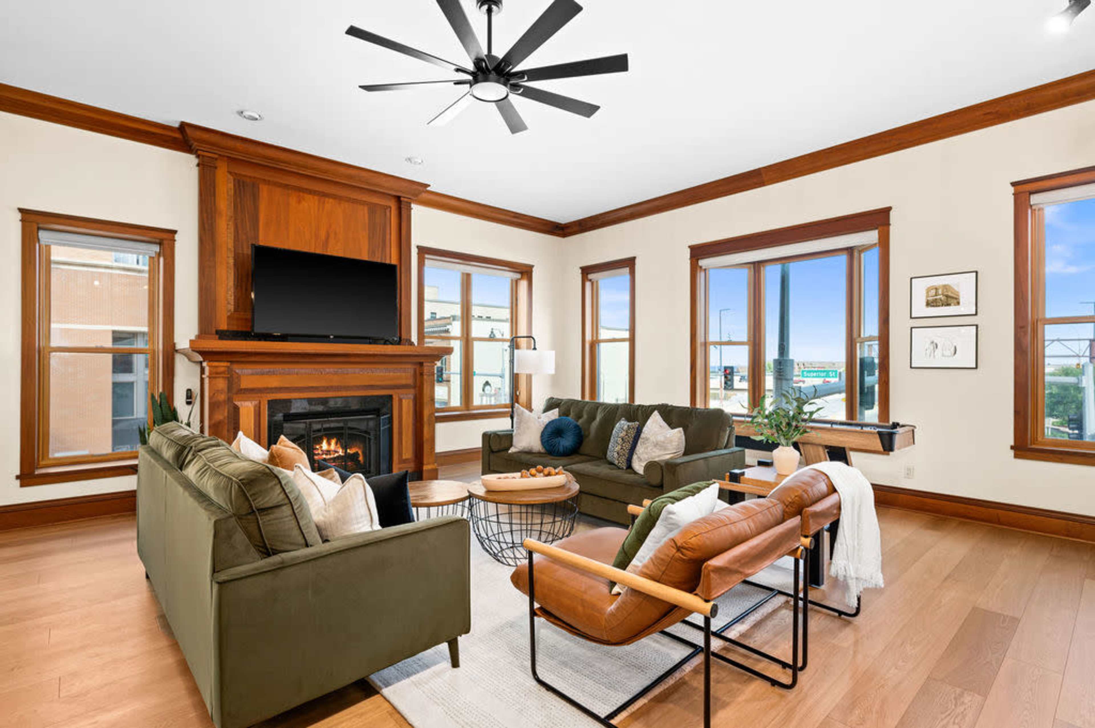 A living room with a green sofa, an armchair, a fireplace with a TV above it, and large windows providing natural light.