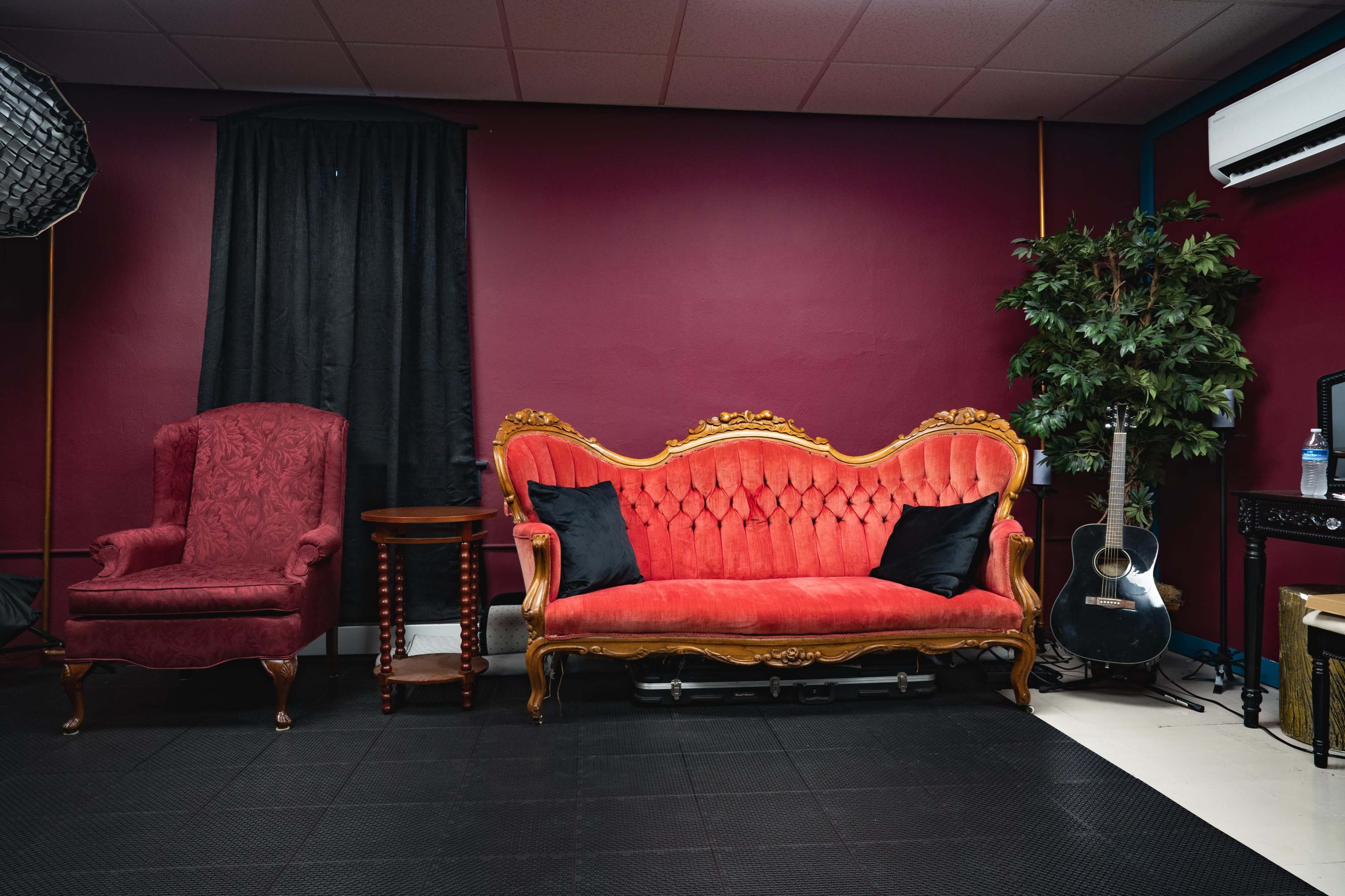 The image shows a lavish red velvet sofa with ornate gold detailing, accompanied by a vintage chair and a guitar, all set against a maroon wall.