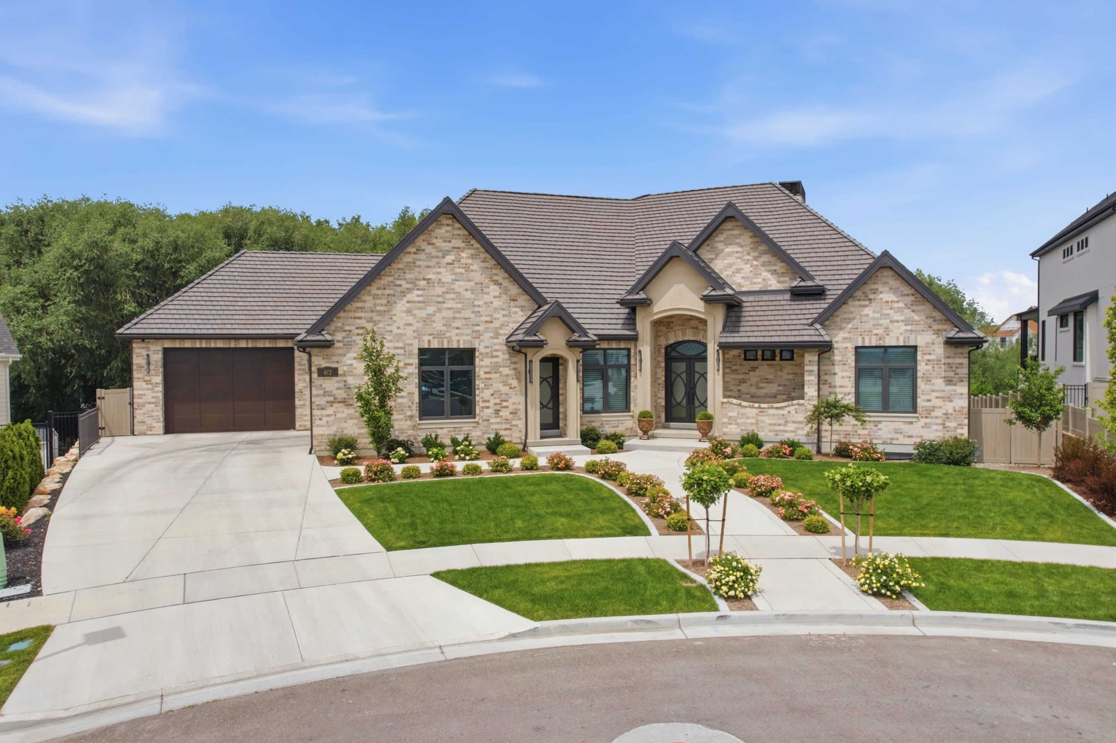 A large, modern stone house with a manicured lawn and circular driveway is situated in a suburban area.