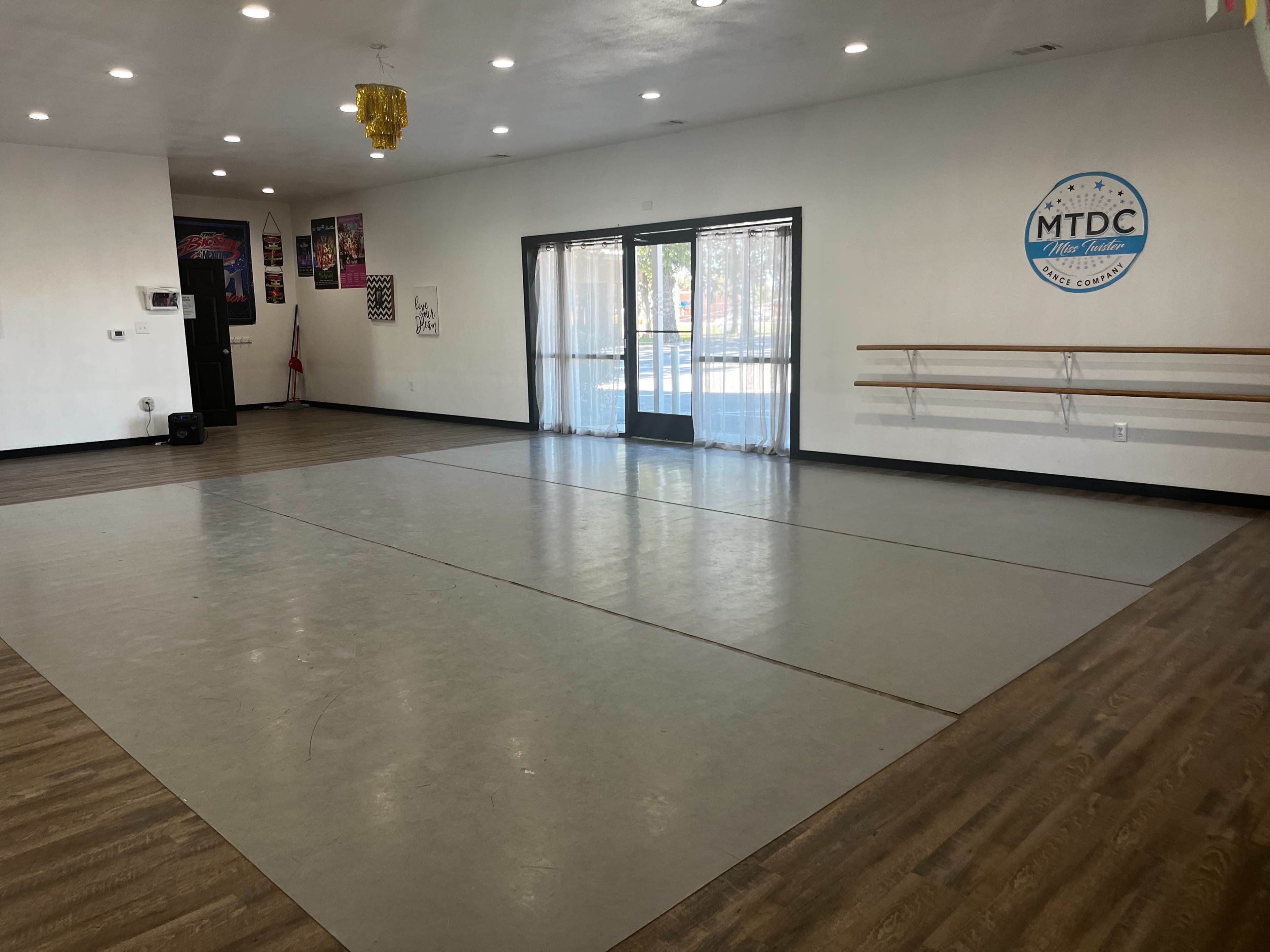 The image shows a spacious dance studio with a light-colored floor, mirrors on one wall, and ballet bars against another.