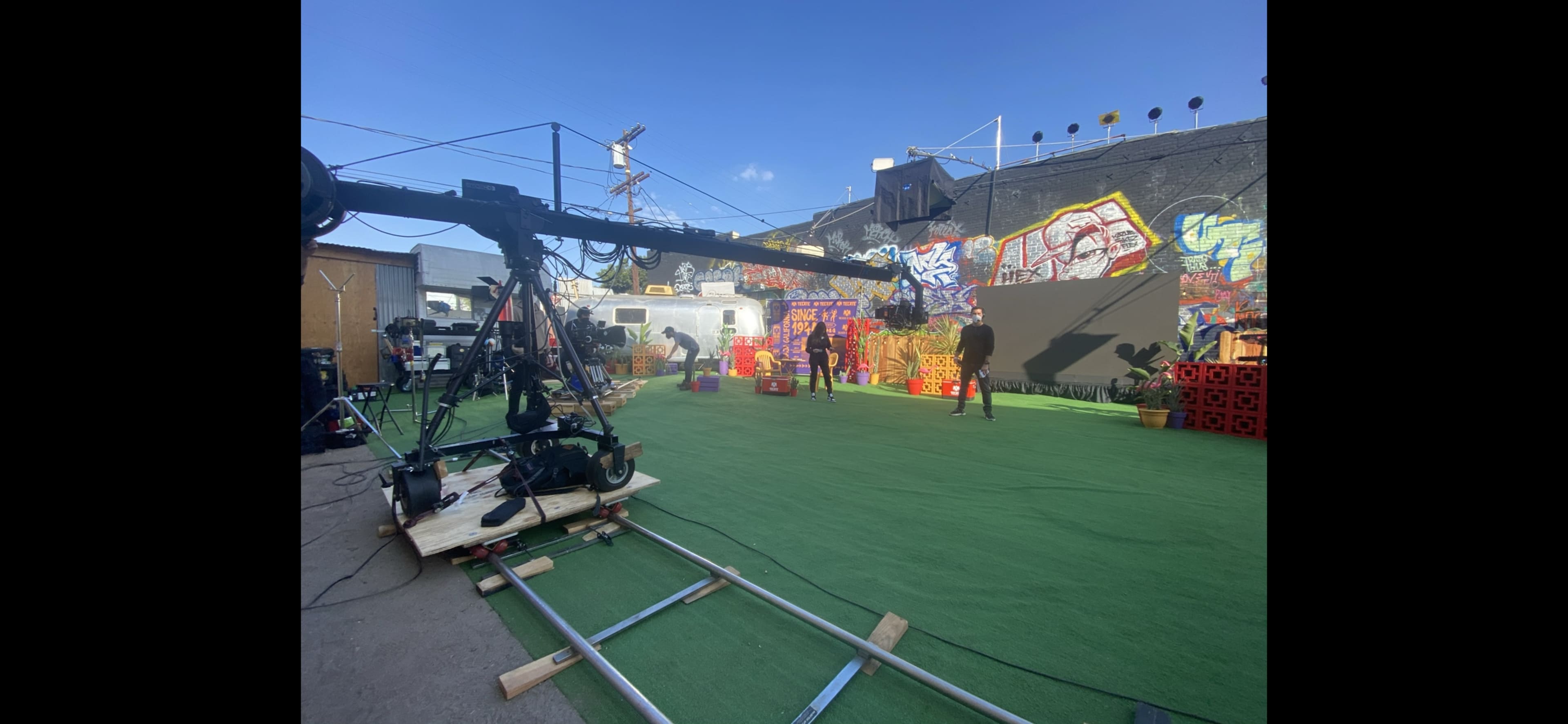 A film set with a camera crane, crew members, and colorful props arranged on a green carpeted area, surrounded by a graffiti-covered wall.
