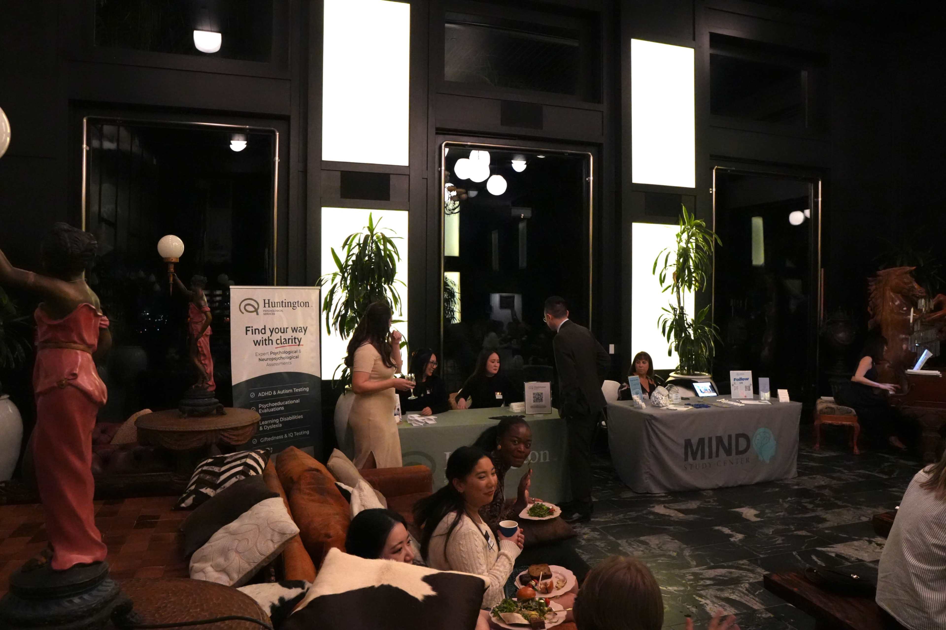 A gathering takes place in a spacious room with large windows and varied seating, featuring attendees interacting at tables while a display for Huntington and MIND is set up in the background.