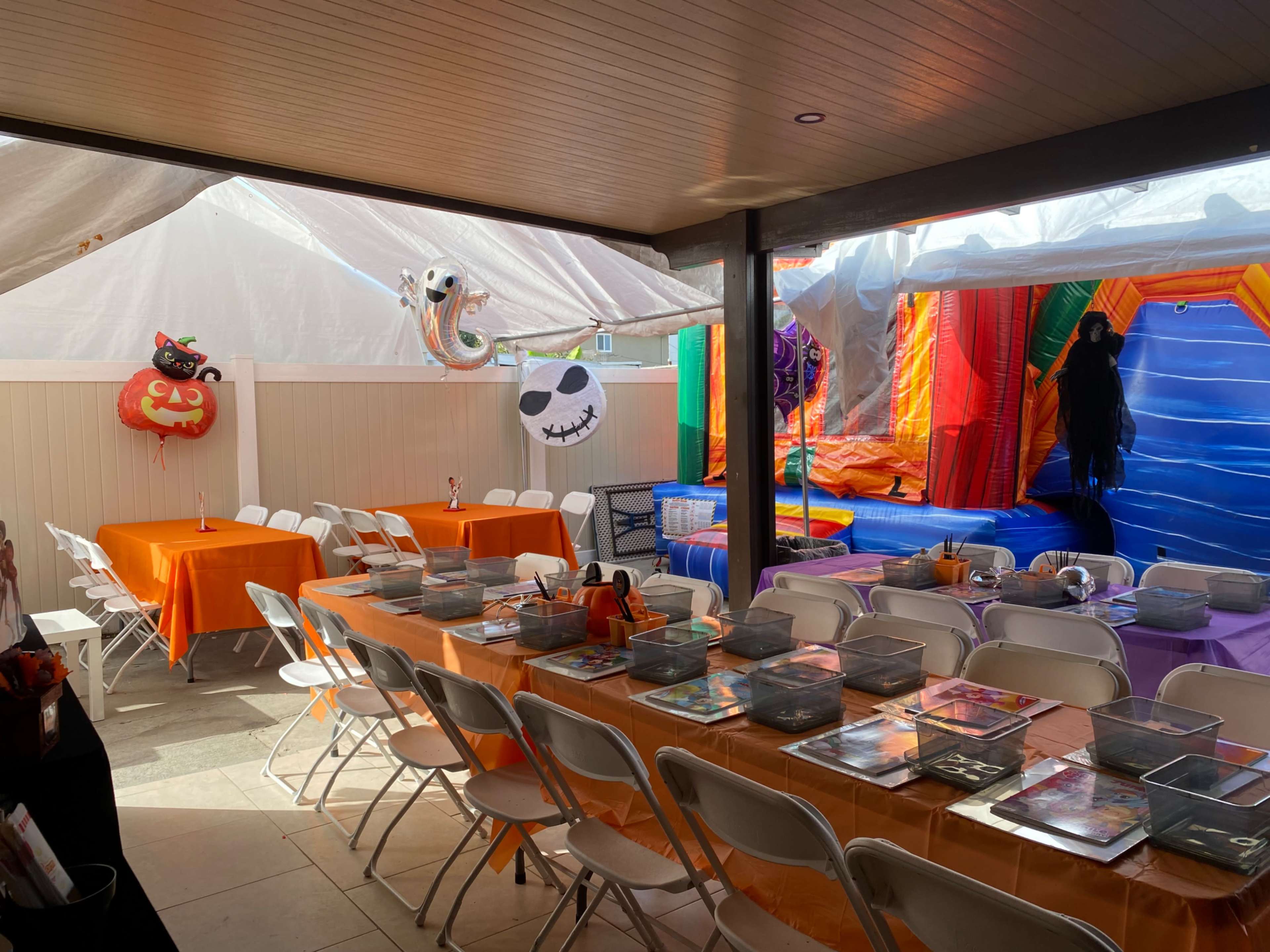 The scene shows a decorated outdoor area for a party with colorful tables, chairs, and a bouncy castle in the background.