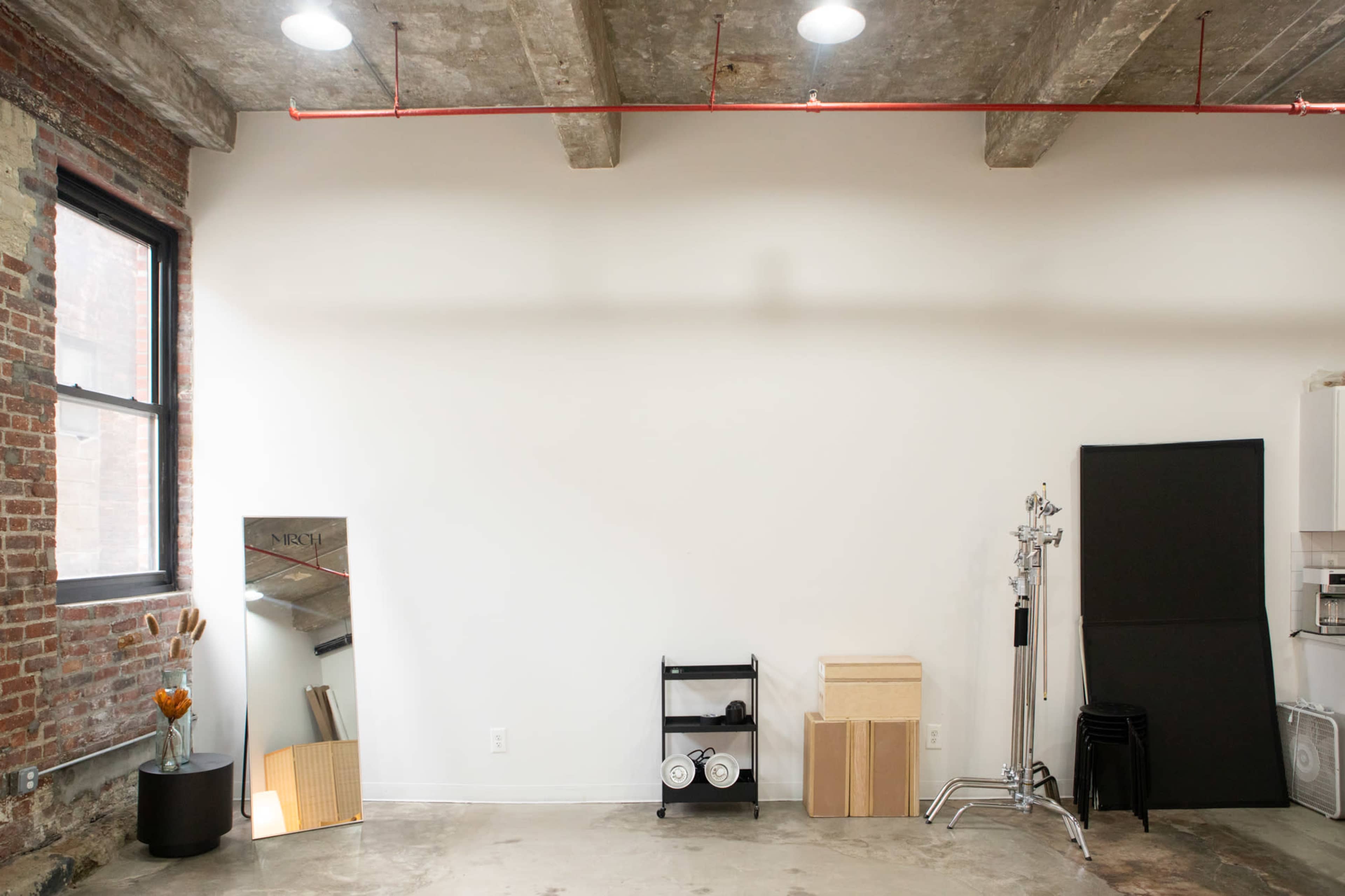 A minimalist interior space featuring exposed brick walls, a large mirror, a small black cart with weights, and stacked wooden boxes against a plain white wall.