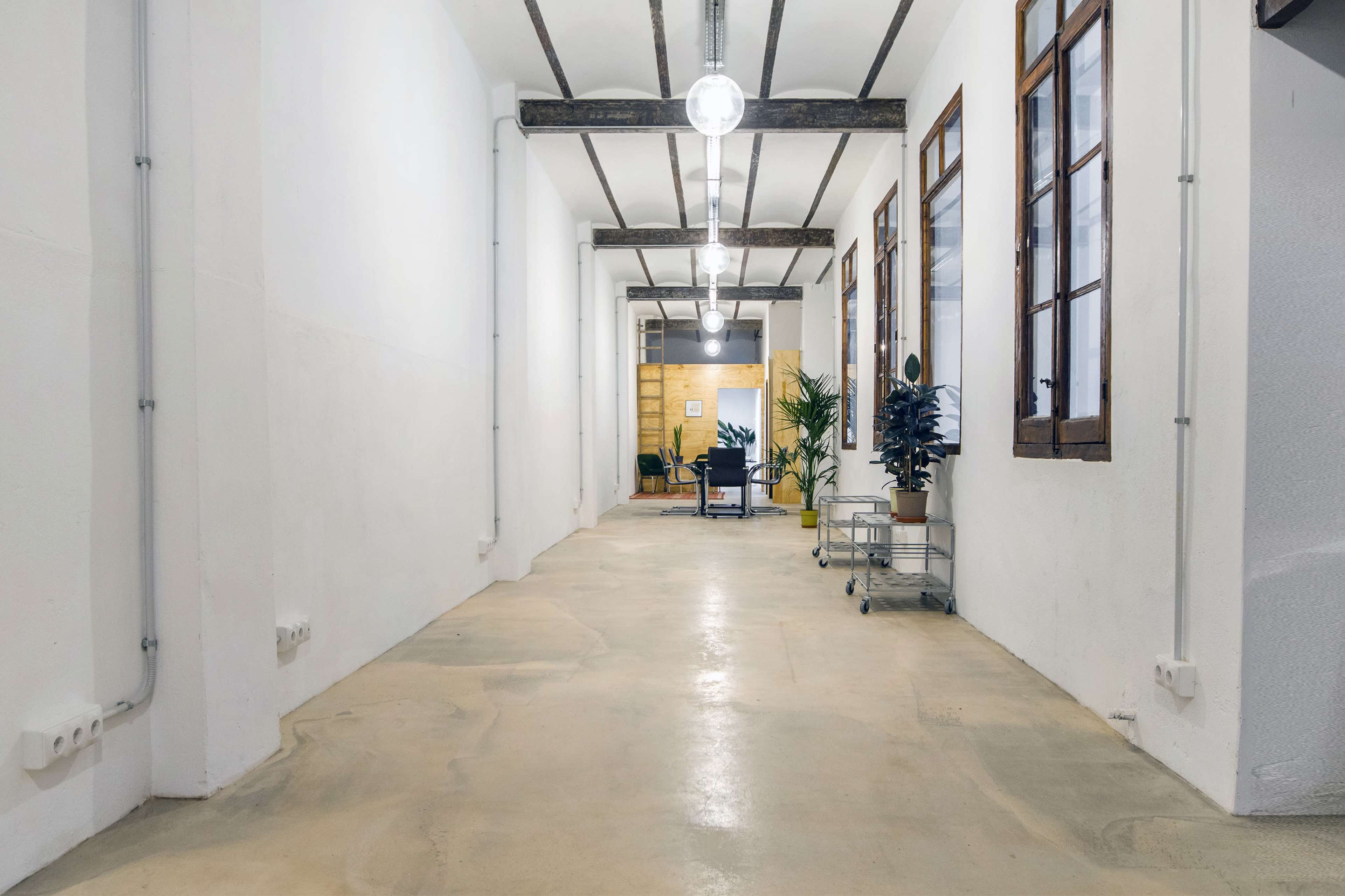 The image shows a long, narrow corridor with white walls, wooden ceiling beams, and windows along one side, leading to a furnished area with plants and furniture at the far end.