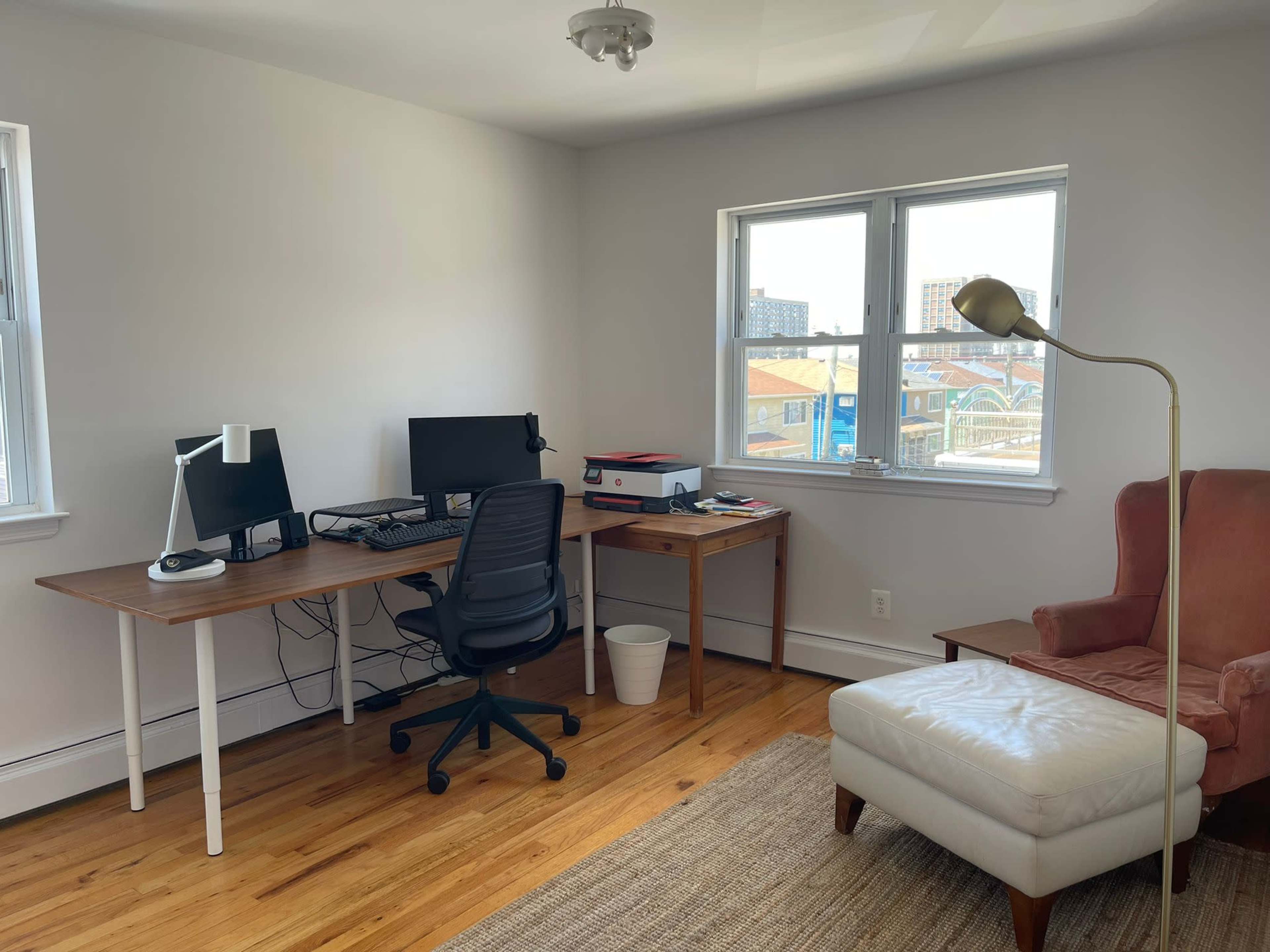 A bright office space with a desk featuring two computer monitors, a chair, and a side table with a lamp and an armchair.