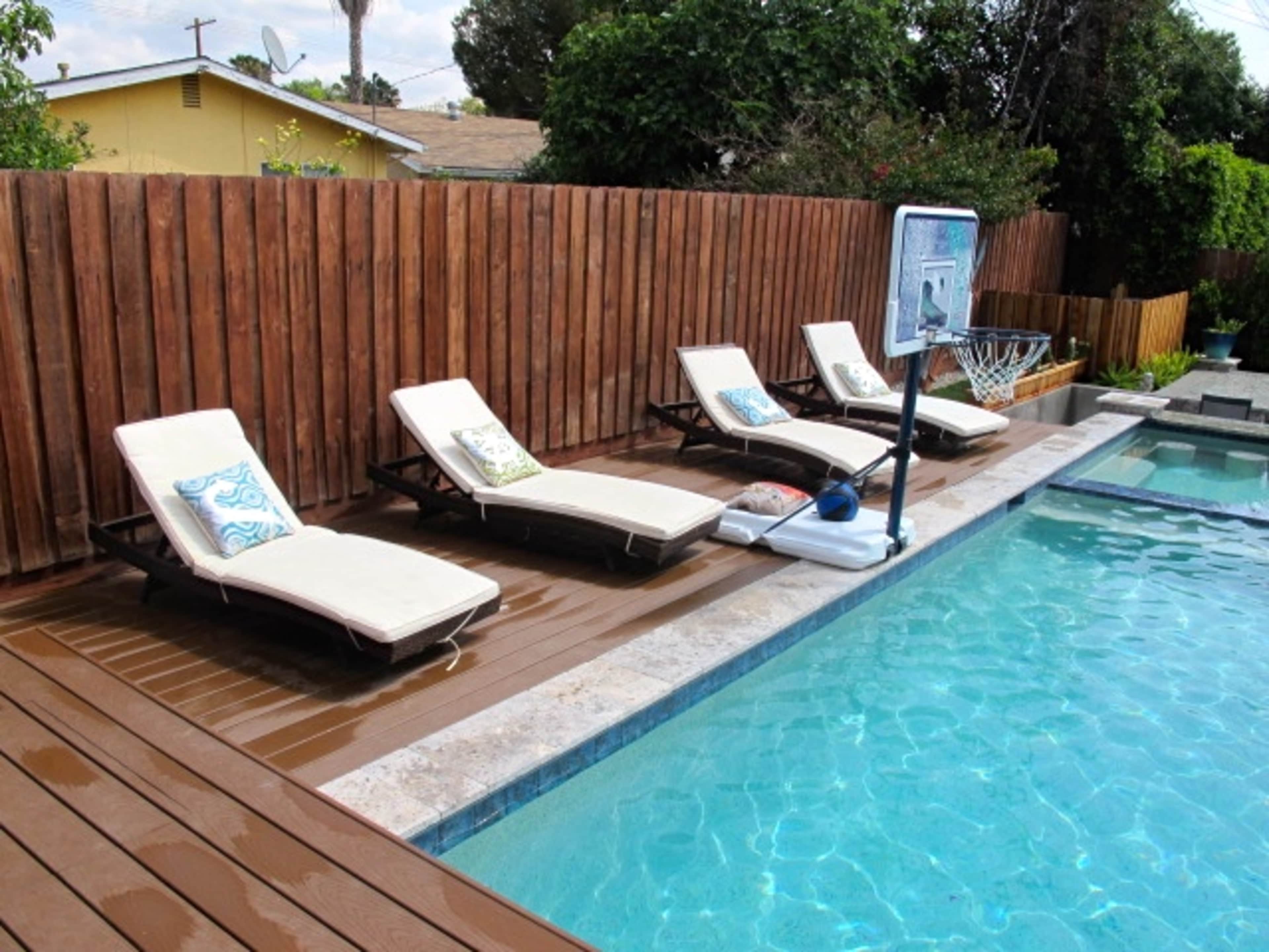 Four lounge chairs with cushions are placed beside a swimming pool, which features a basketball hoop nearby, all enclosed by a wooden fence.