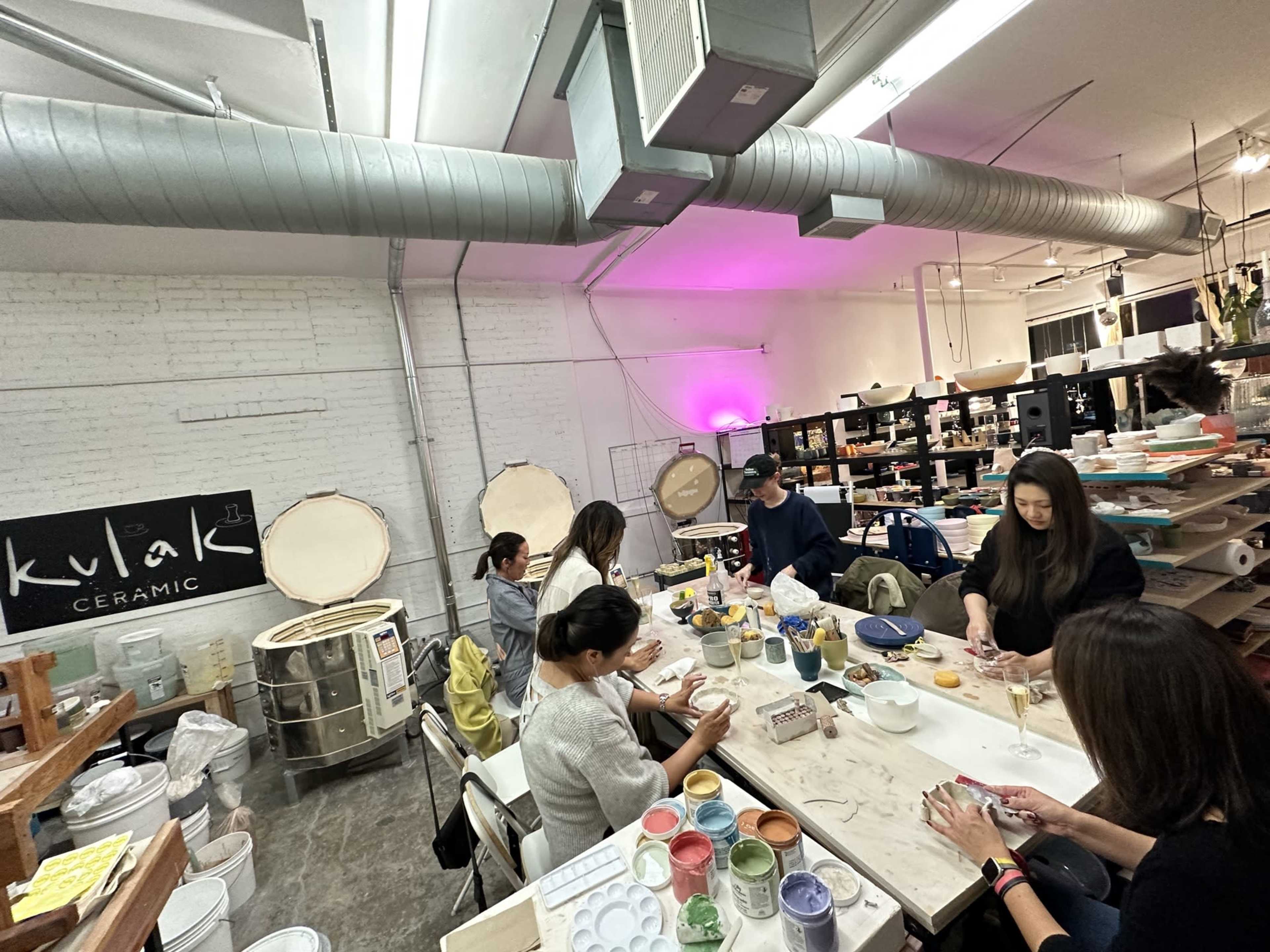 A group of people is seated at a table in a ceramics studio, engaged in pottery-making activities while various tools and materials are organized around them.