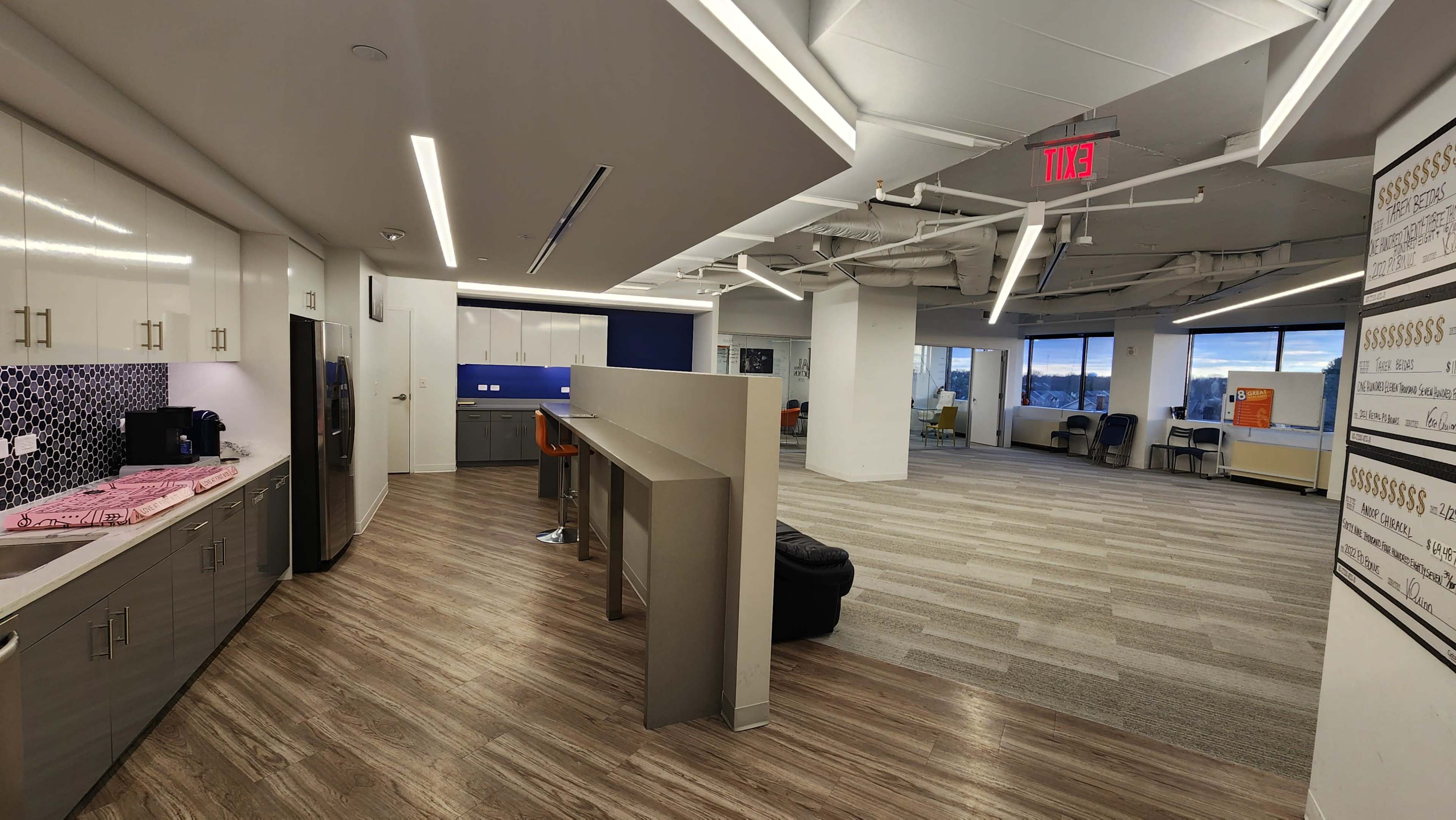 A modern office space featuring a kitchen area with appliances and seating, an open workspace with large windows, and a carpeted floor.