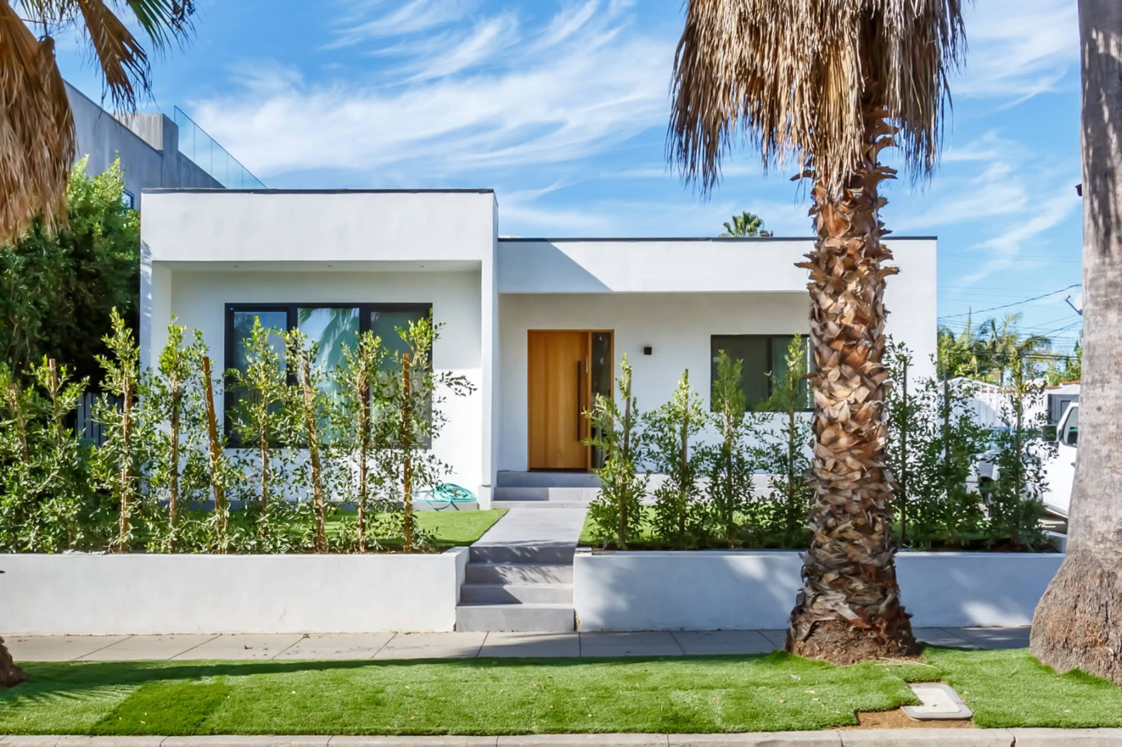 A modern white house with a wooden door is surrounded by green landscaping and palm trees.