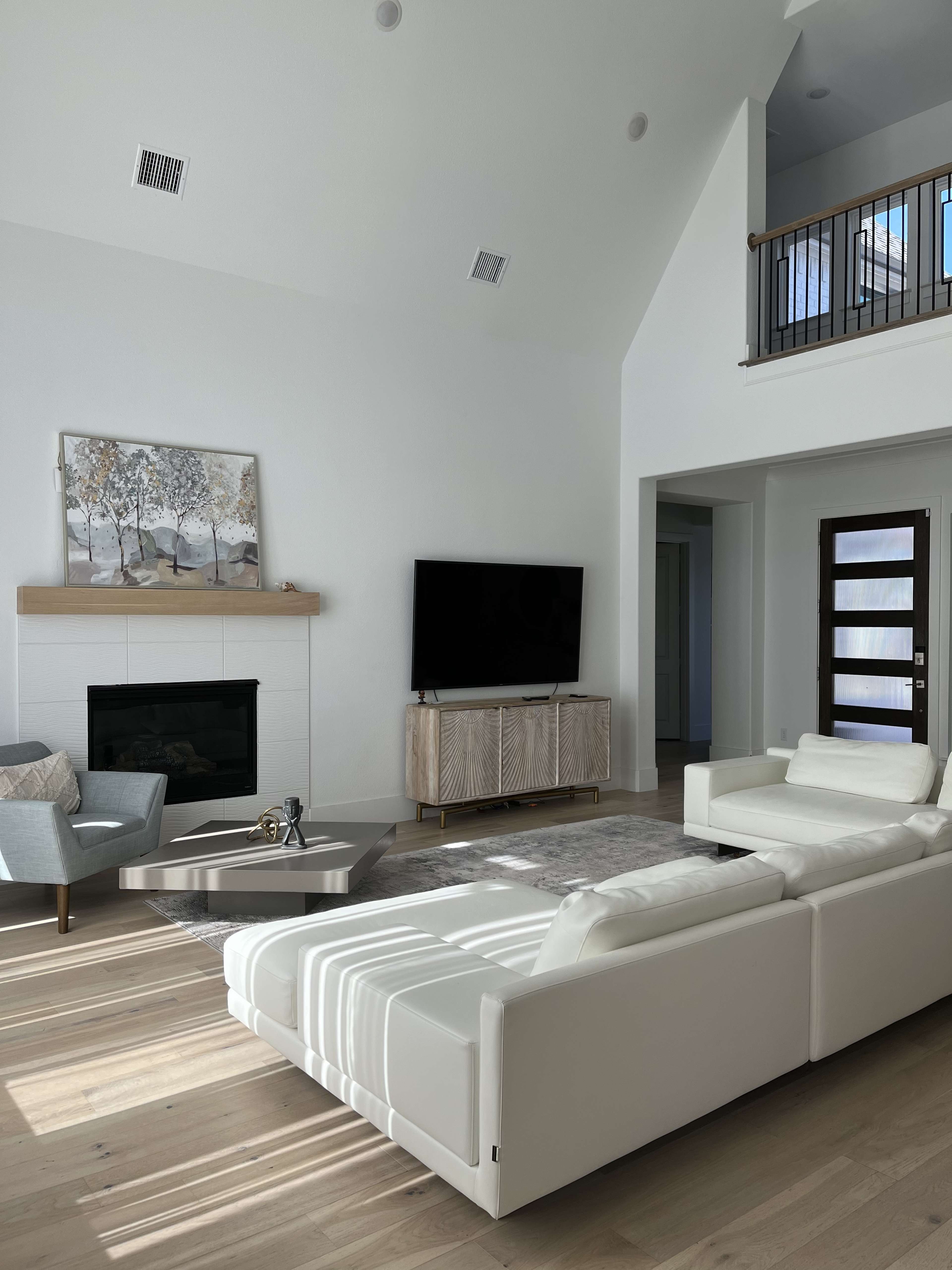 The image shows a modern living room featuring a large white sectional sofa, a glass coffee table, a TV mounted on the wall, and a staircase leading to a balcony.