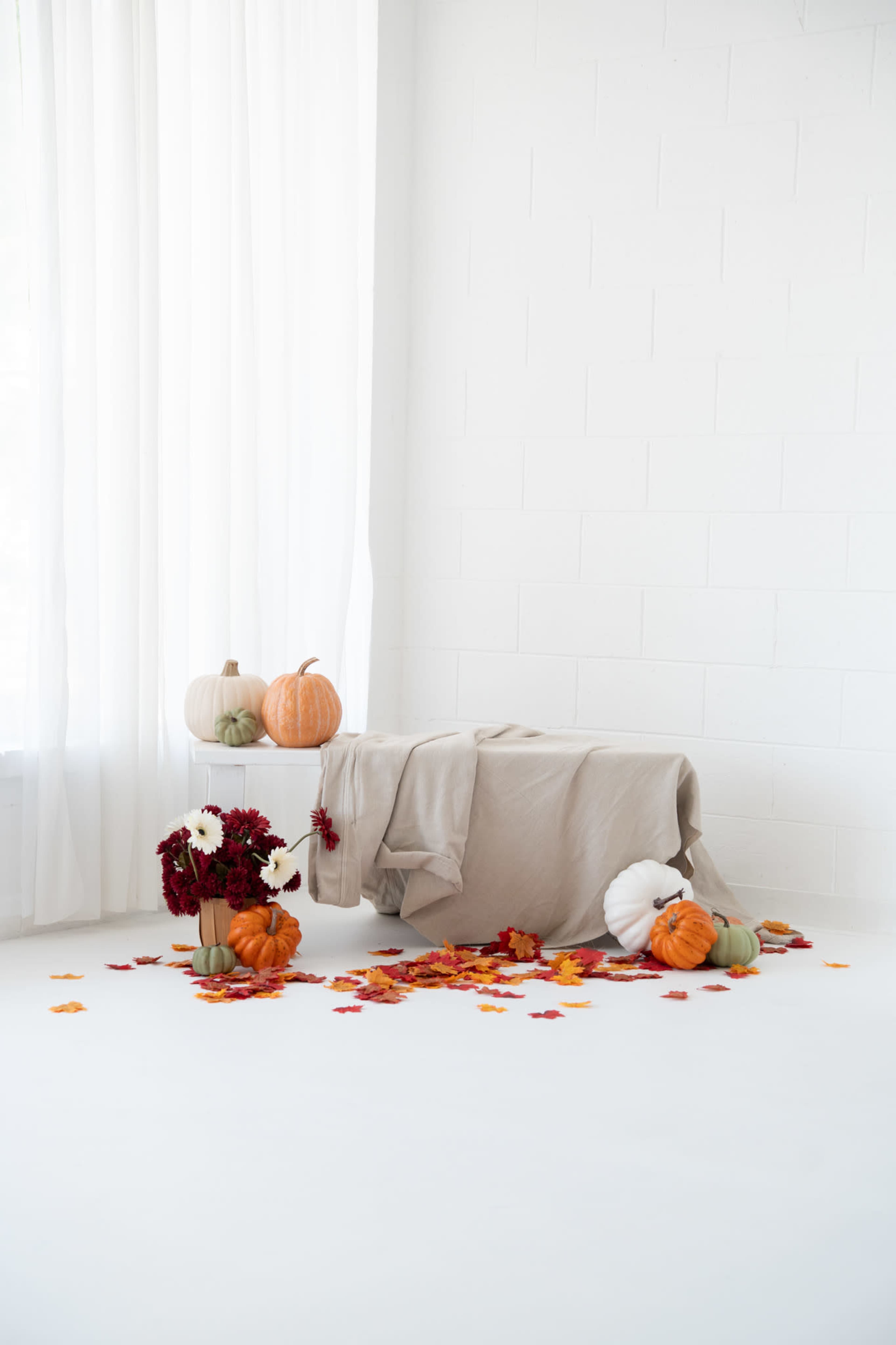 A beige tablecloth drapes over a table adorned with pumpkins and floral arrangements amidst scattered autumn leaves on a white floor.