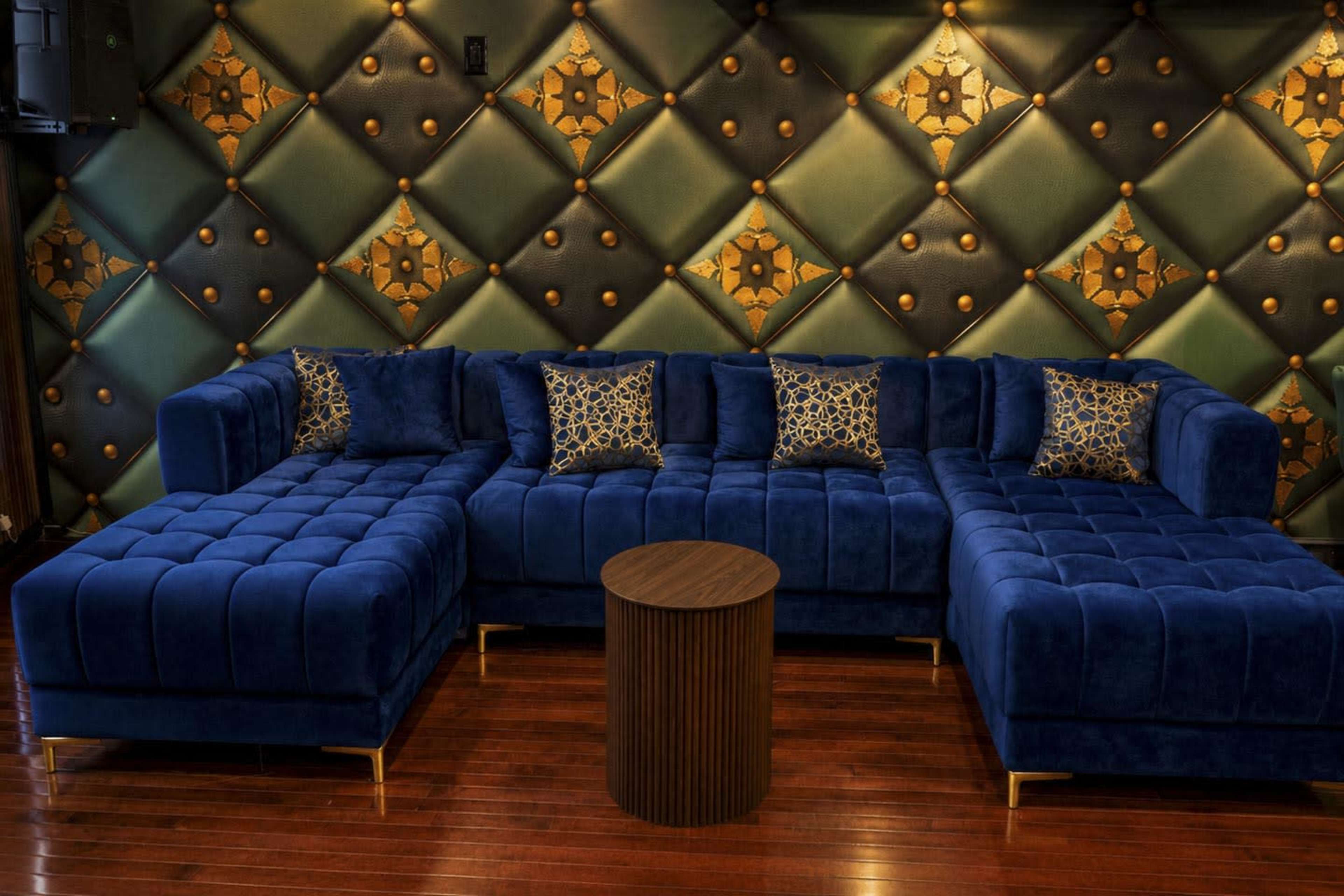 The image shows a blue tufted sectional sofa with decorative pillows, set against a patterned wall with green and gold designs and a round wooden coffee table in the center.