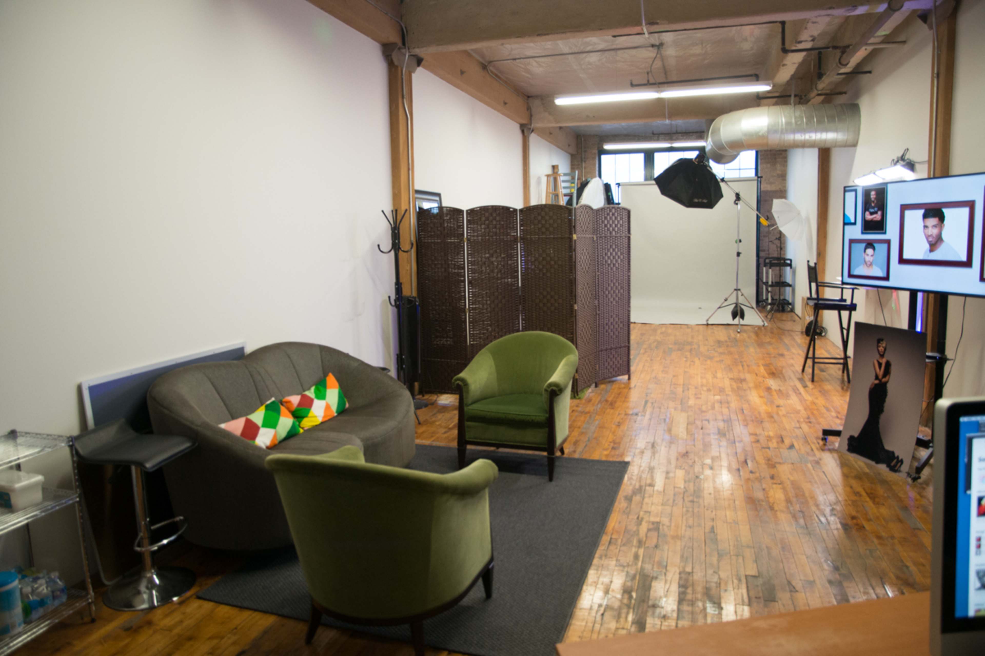 A spacious studio interior with two green chairs and a gray sofa against a wooden floor, partitioned by a folding screen, and equipped with photography equipment in the background.