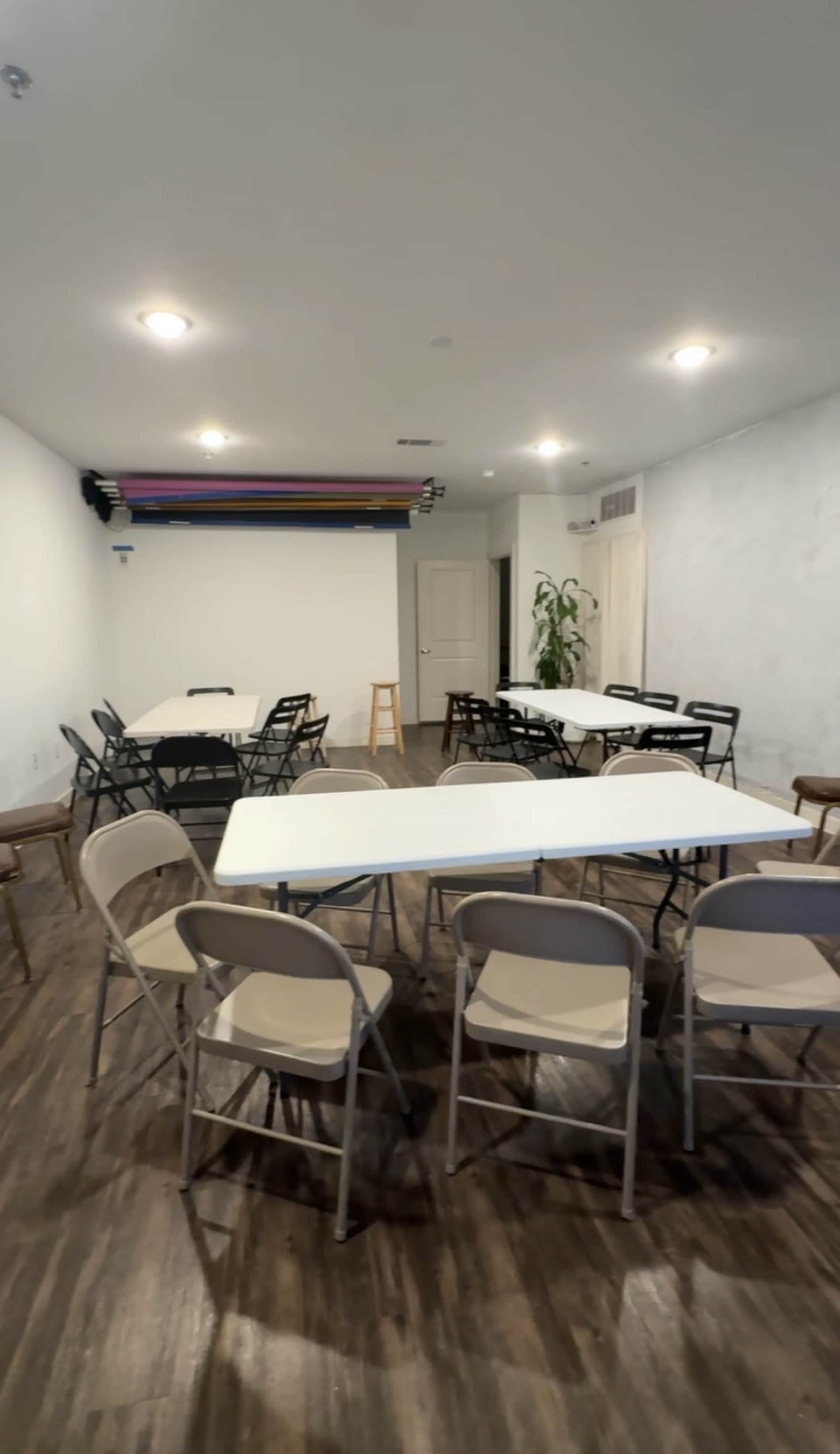 The image shows a spacious room set up with several white tables and folding chairs arranged around the space, with a backdrop system mounted on the wall.