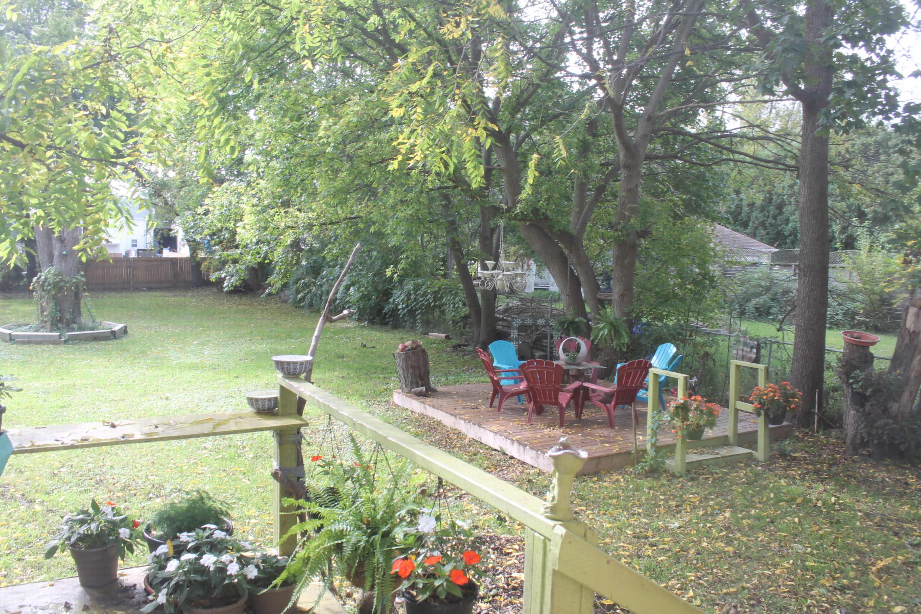 A spacious backyard with a patio area featuring colorful chairs surrounded by trees and garden plants.