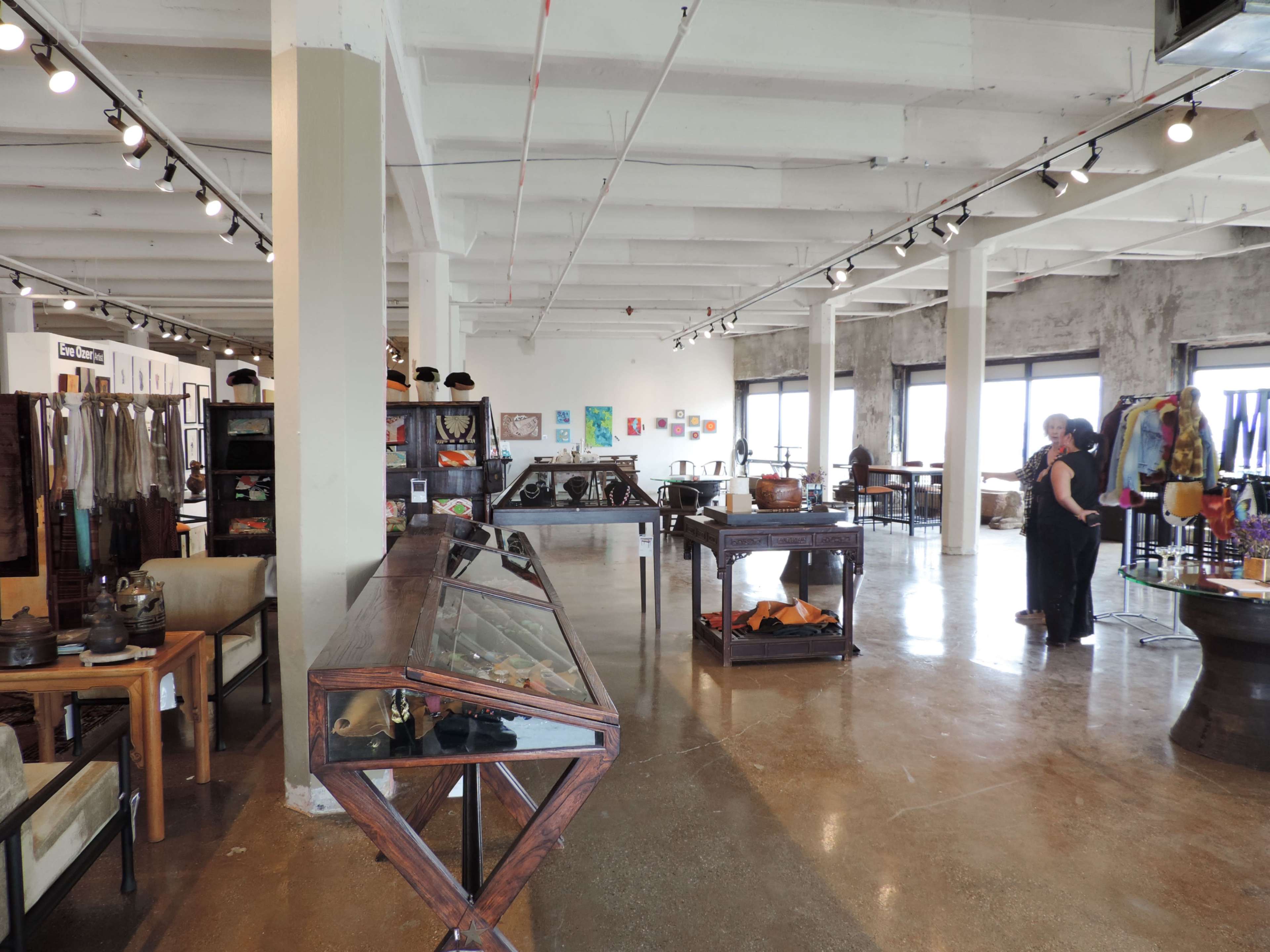 The image shows a spacious, industrial-style art gallery or boutique with various displays of art and merchandise, featuring an open floor plan and large windows.