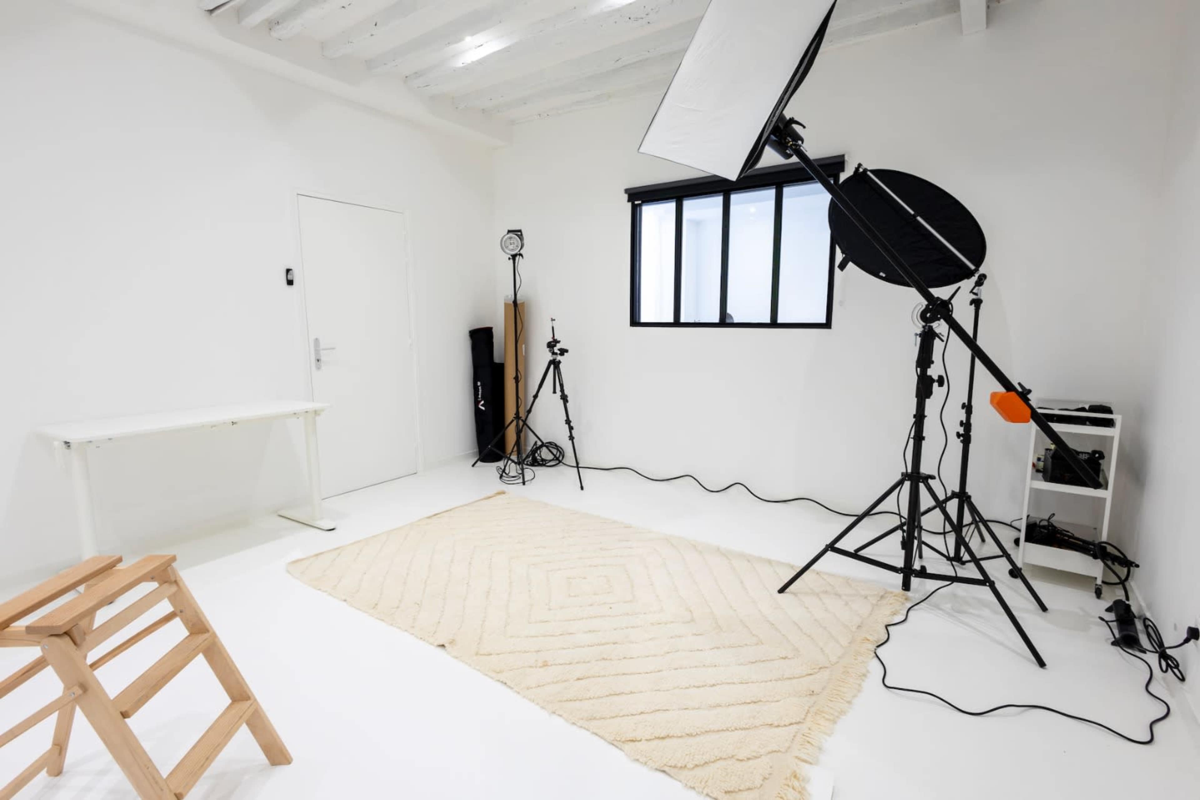 The image shows a minimalist photography studio with white walls, a large window, a light stand, and a woven rug on the floor.